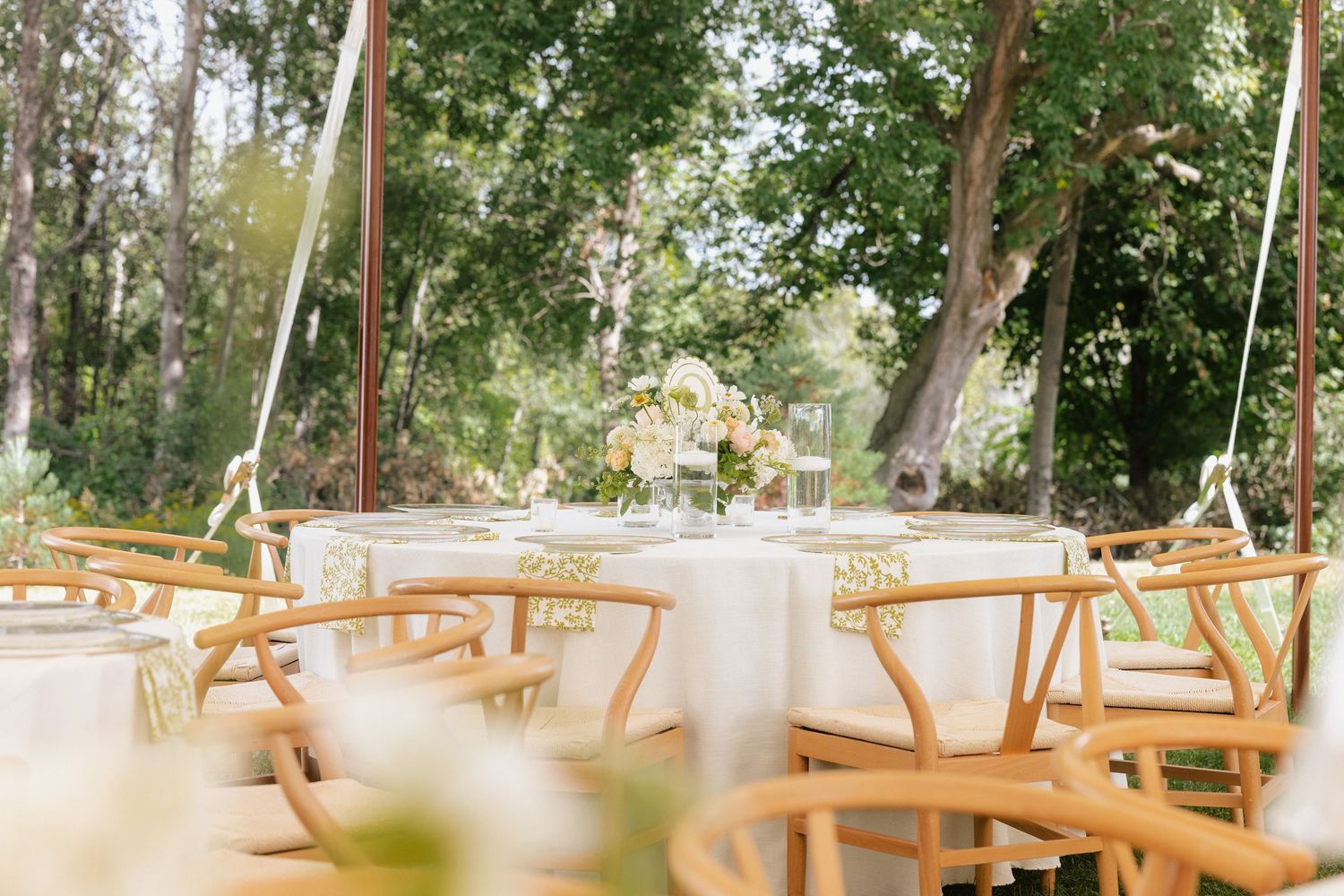 Elegant outdoor wedding reception with wooden chairs and white tablecloths under a canopy surrounded by lush green trees.