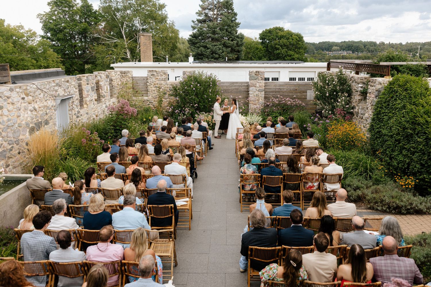 Outdoor wedding ceremony in a walled garden with guests seated in wooden chairs facing the altar.