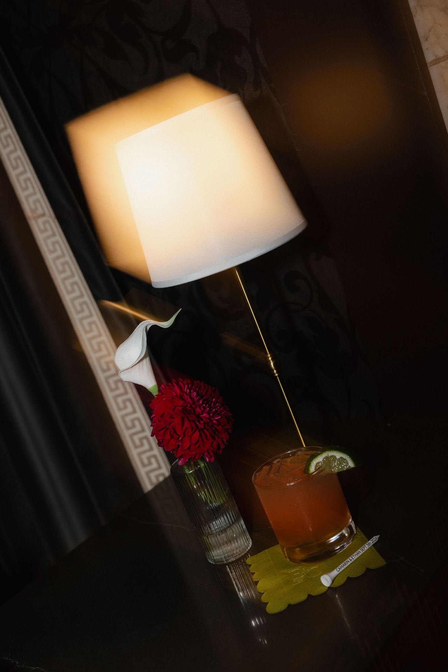 Dimly lit cocktail table with red flowers, white lamp, and whiskey drink garnished with lime slice.
