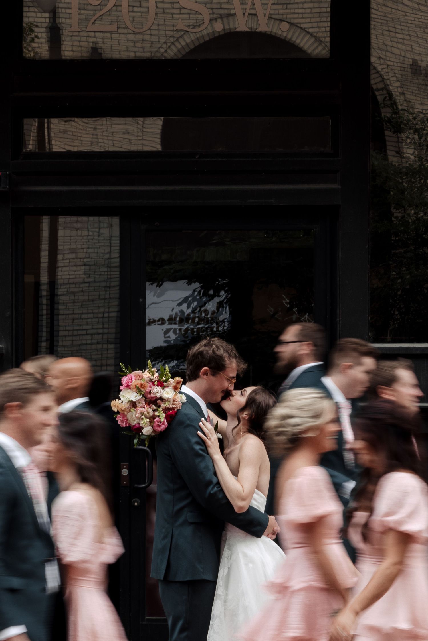 Bride and groom kissing with colorful bouquet as wedding party moves around them in motion blur.