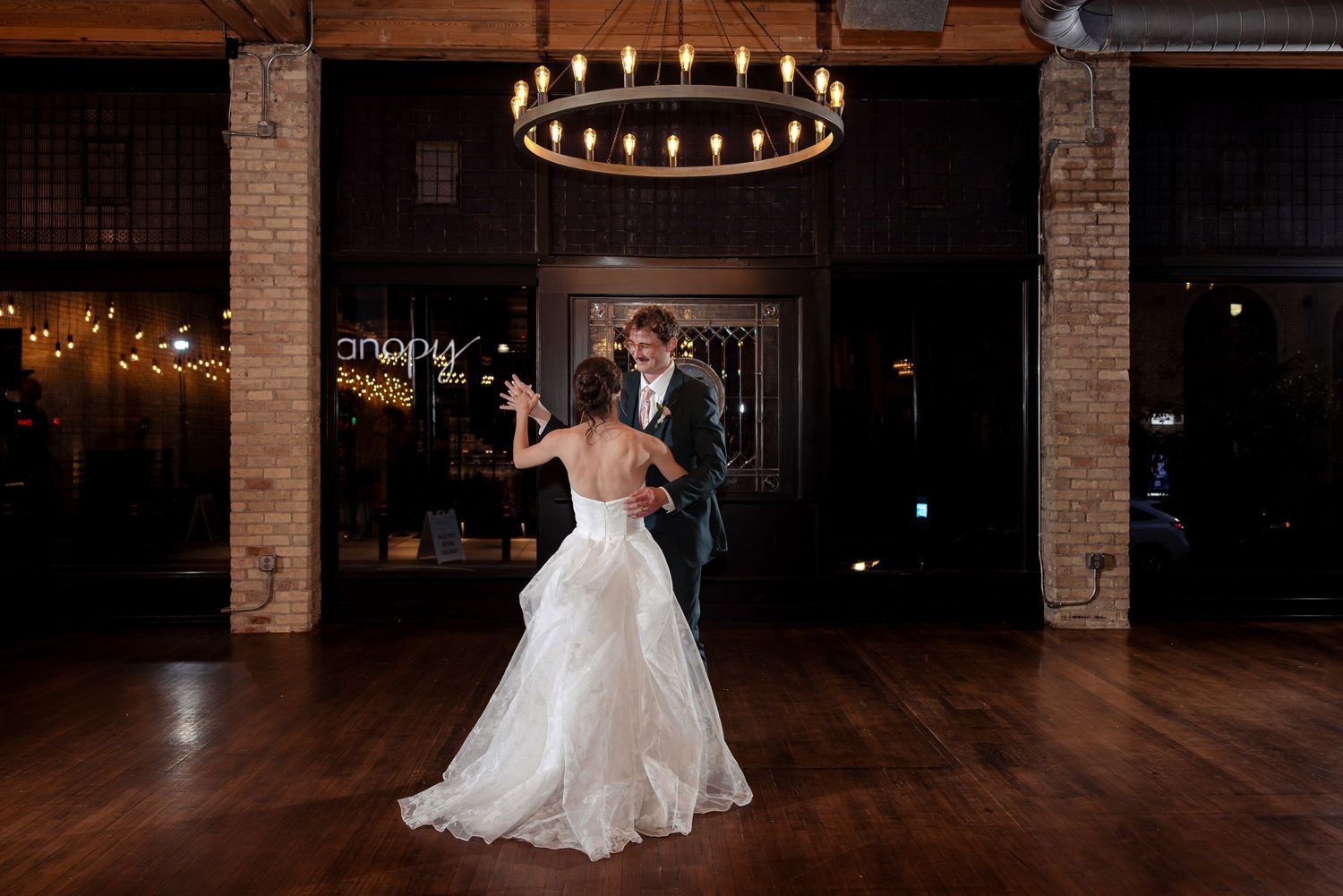 Bride and groom sharing their first dance under a circular chandelier on a hardwood floor in a brick venue.