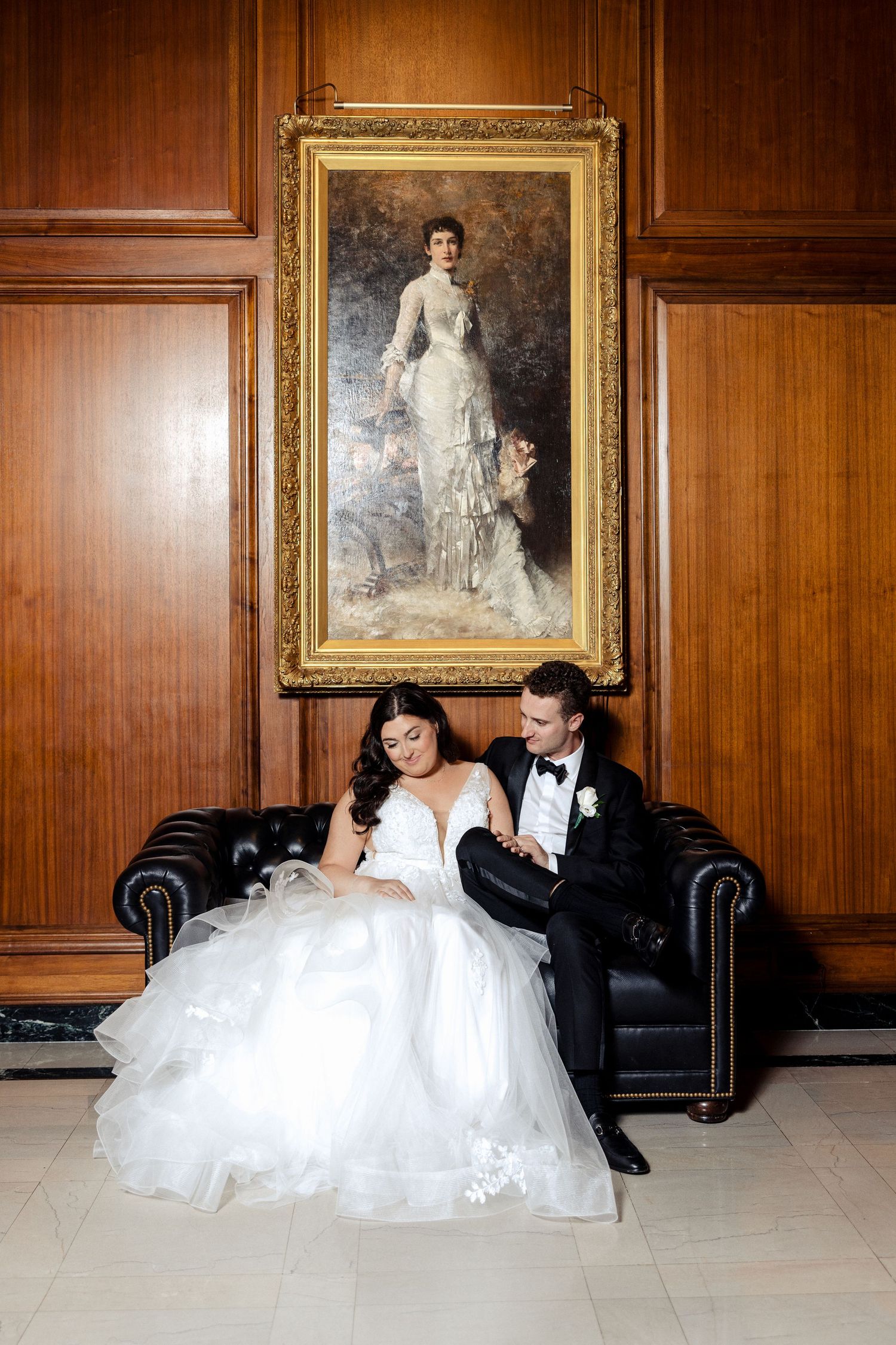 Bride and groom seated on a leather sofa beneath a golden-framed portrait in a wood-paneled room.