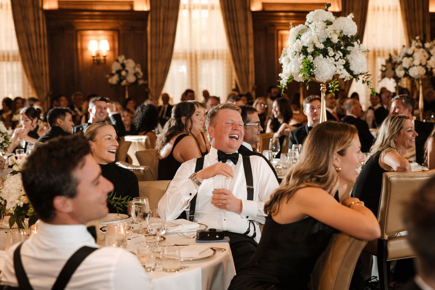 Wedding guests laughing and enjoying speeches at an elegant floral-decorated reception dinner.