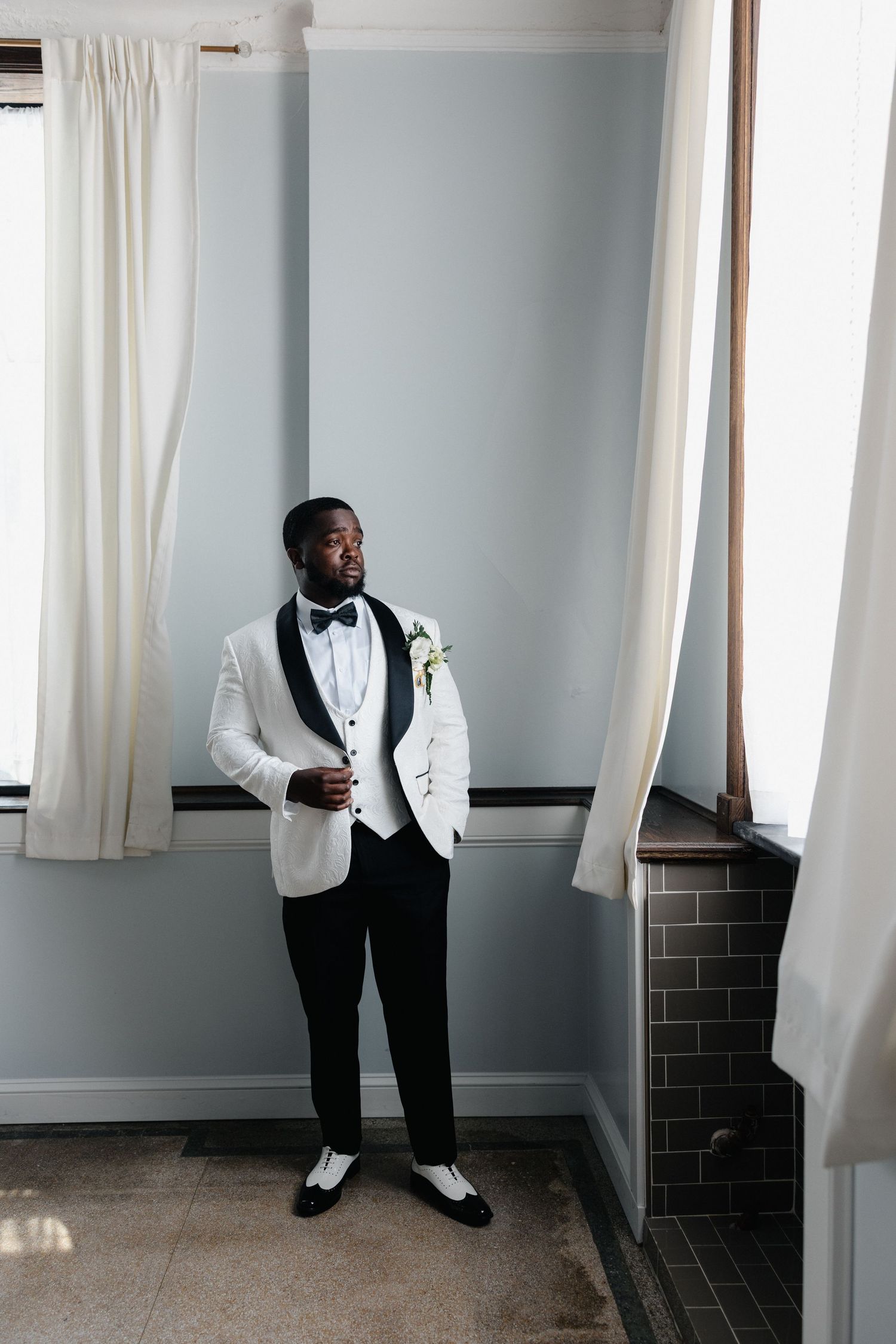 Groom in white tuxedo jacket with black lapels and boutonniere stands confidently by window in elegant room.
