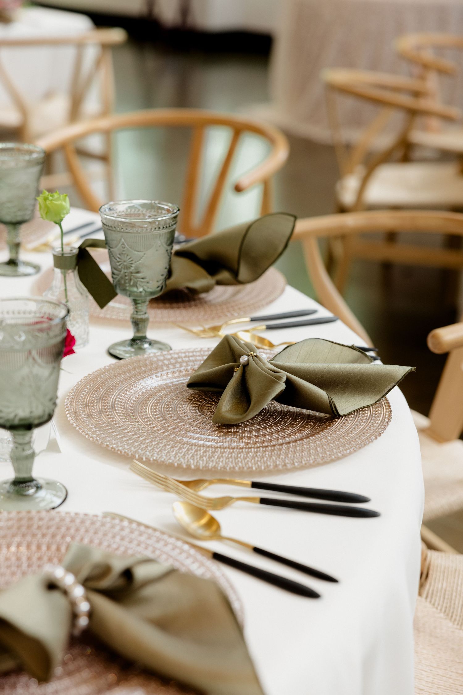 Elegant wedding table setting with sage green napkins, gold cutlery, pink charger plates, and green glassware on white linen.