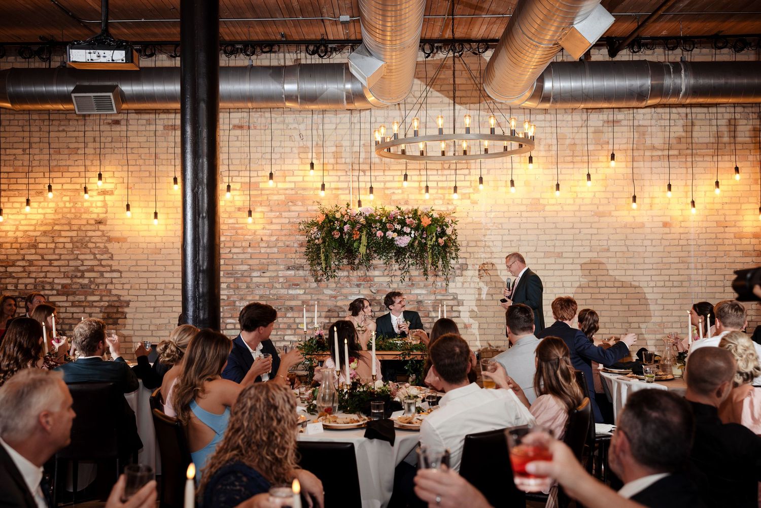 Wedding reception dinner scene with guests seated at round tables under Edison bulb lights in a brick-walled venue.