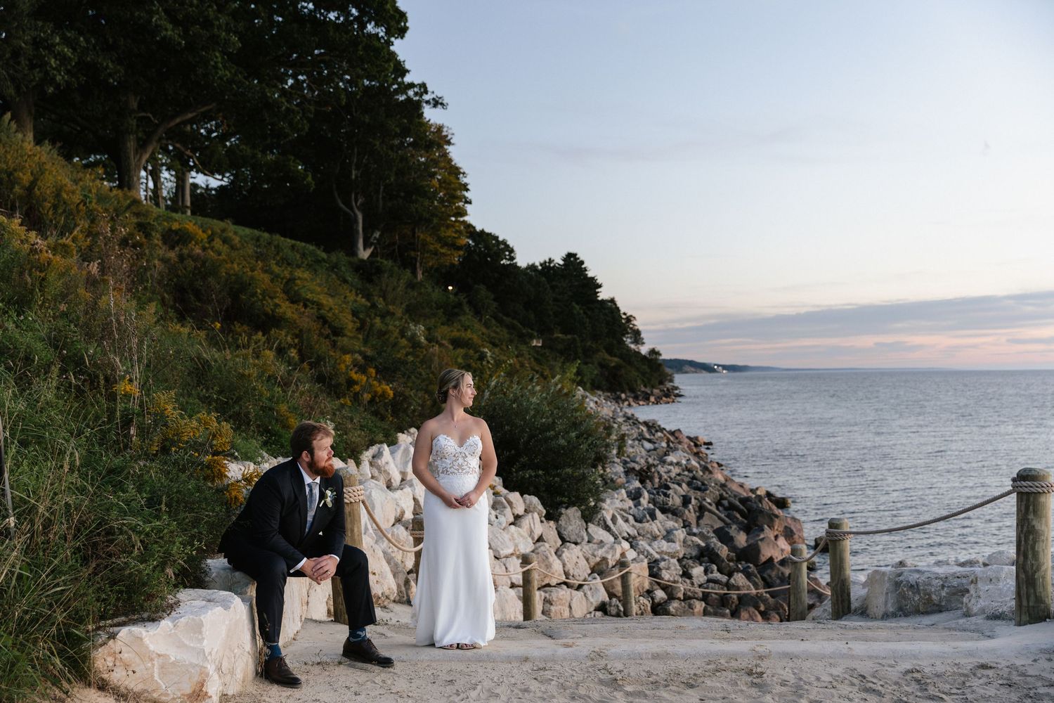Bride in white gown and groom in navy suit pose by rocky shoreline at sunset with trees in background.