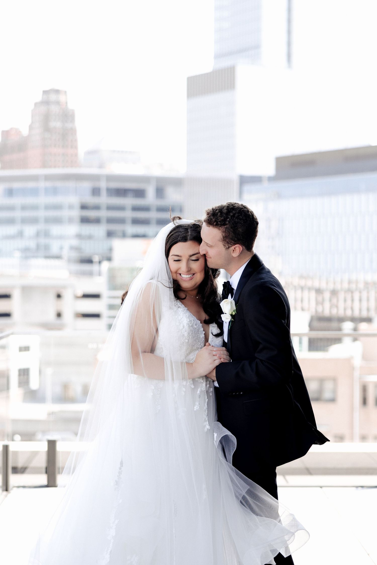 Groom whispers to smiling bride on a rooftop, she wears a lace wedding gown and long veil with city views.