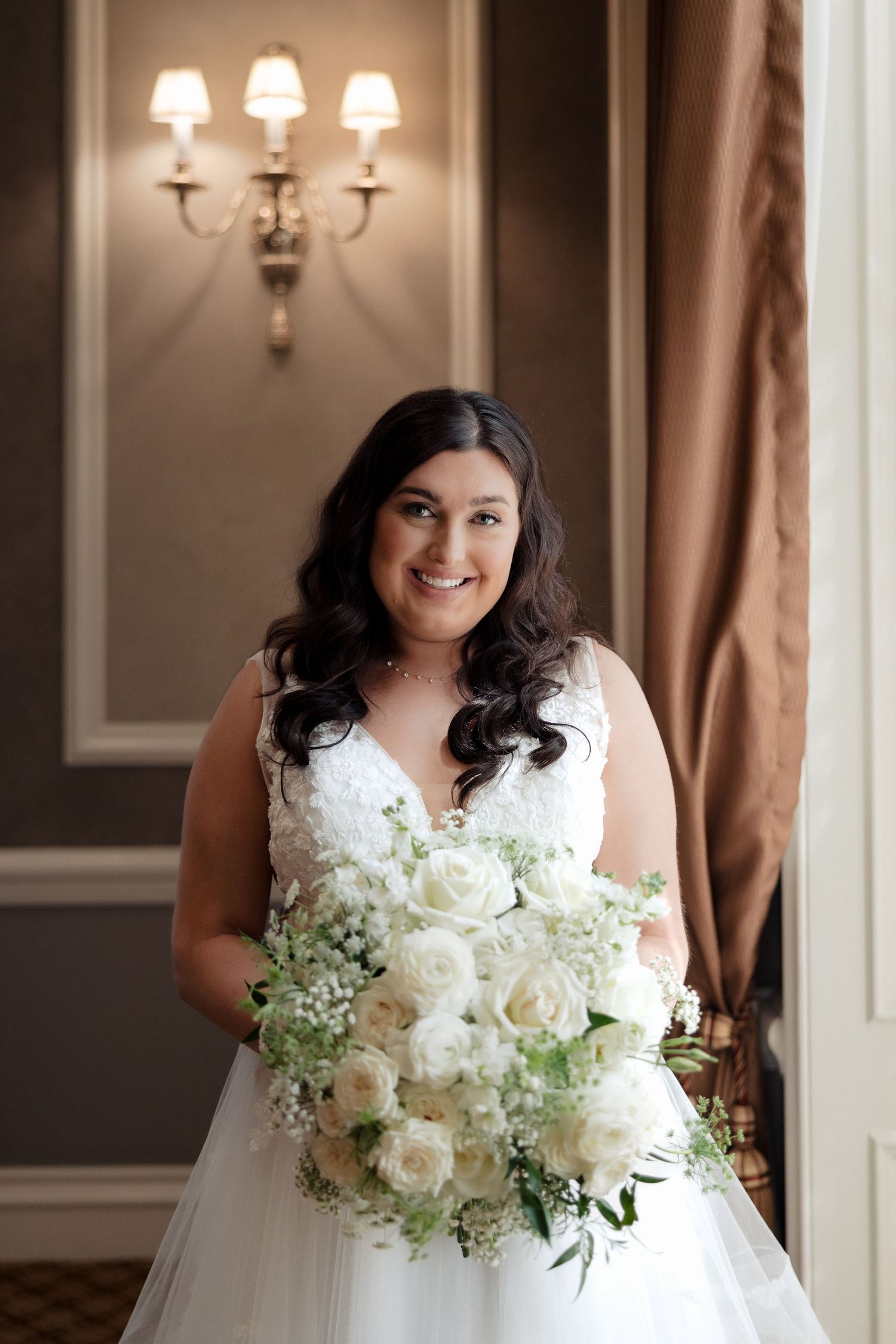 Radiant bride in white gown smiles holding white rose bouquet indoors.