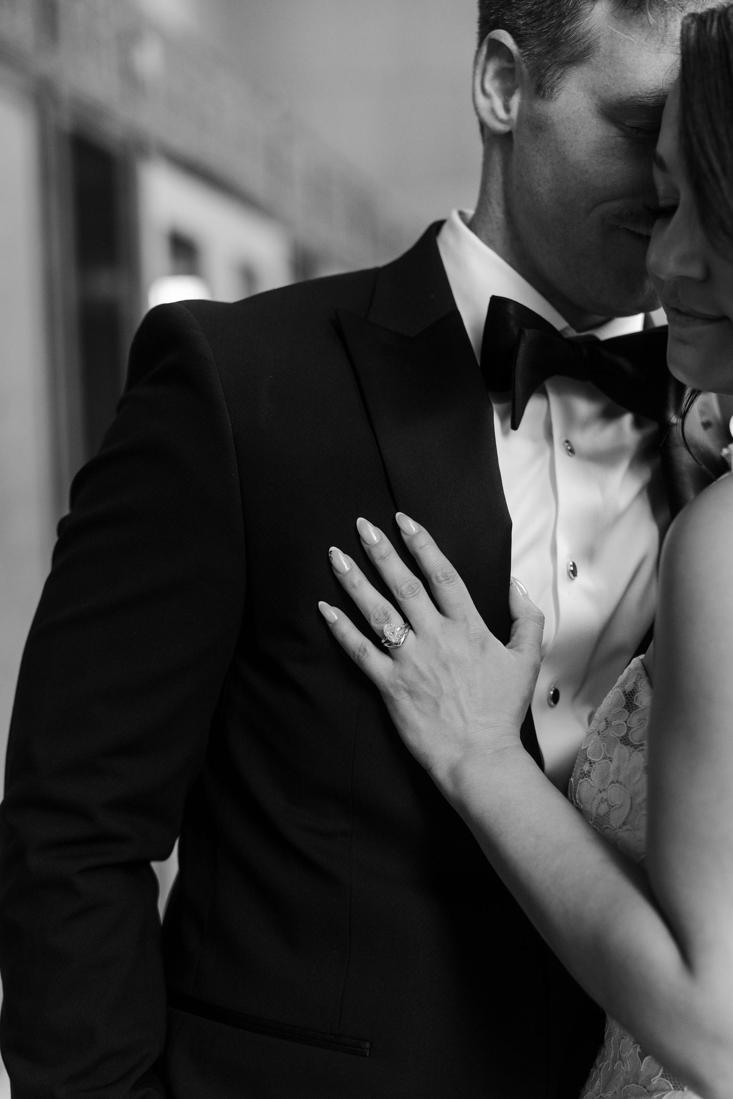 Black and white wedding photo of bride placing hand on groom's tuxedo chest, showcasing elegant diamond engagement ring.