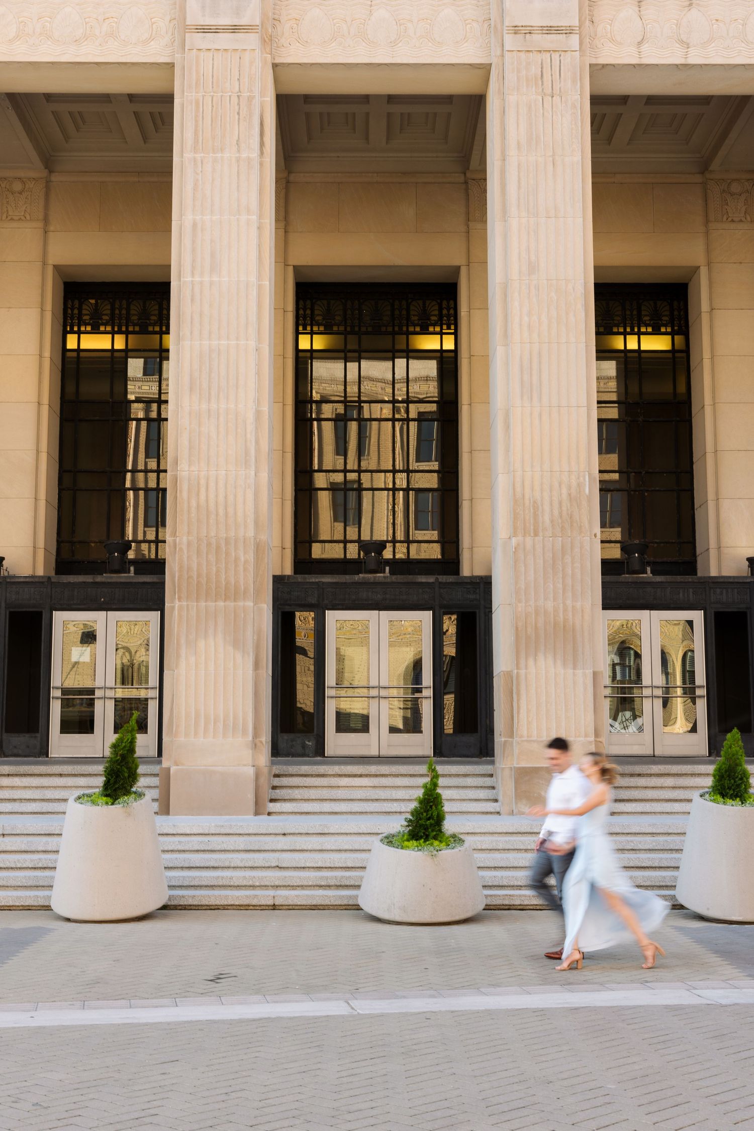Two blurred pedestrians walk past a grand neoclassical building facade with tall columns and ornate entrance doors.