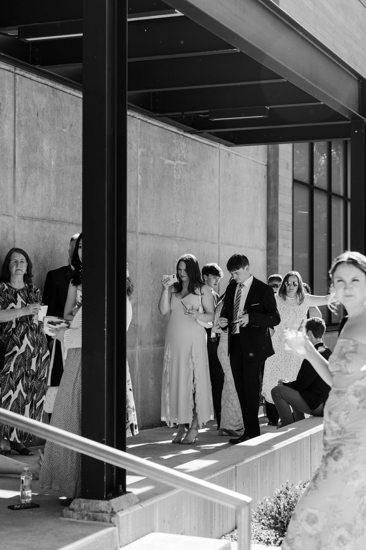 Black and white photo of elegantly dressed wedding guests socializing outside a modern concrete building.