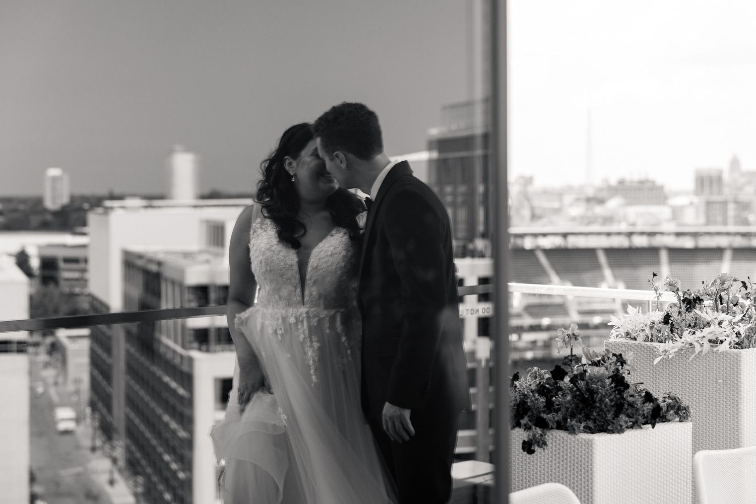 Black and white photo of a bride and groom kissing by a window with a city skyline behind them.