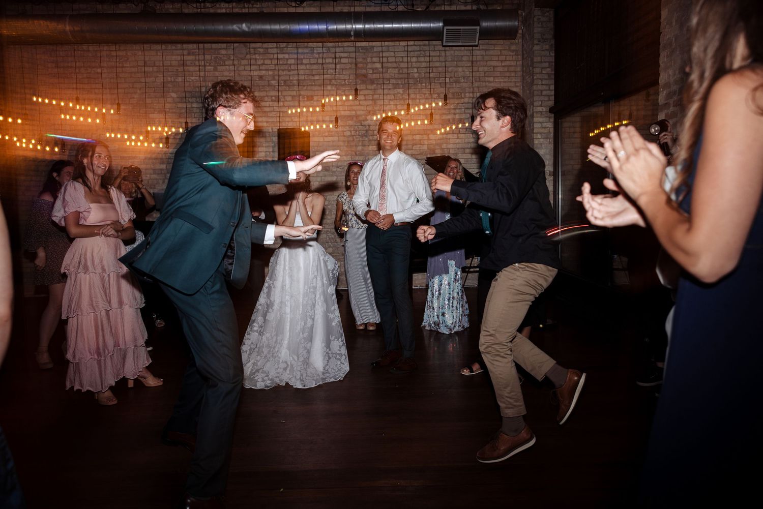 Guests celebrate on the dance floor at a wedding reception in a brick venue with string lights.