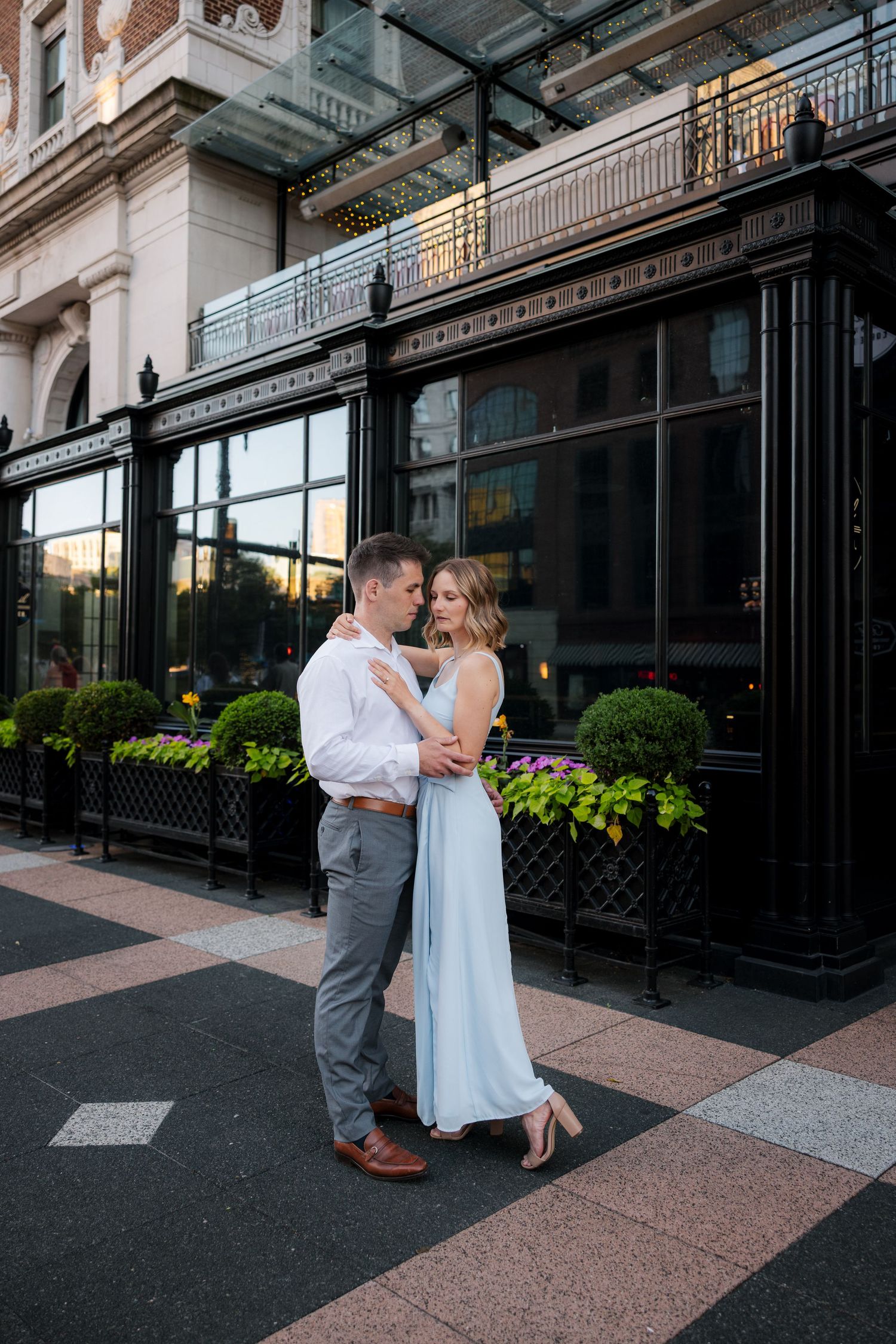 Couple embracing in light blue dress and white shirt outside elegant dark-windowed building with flower boxes at golden hour.