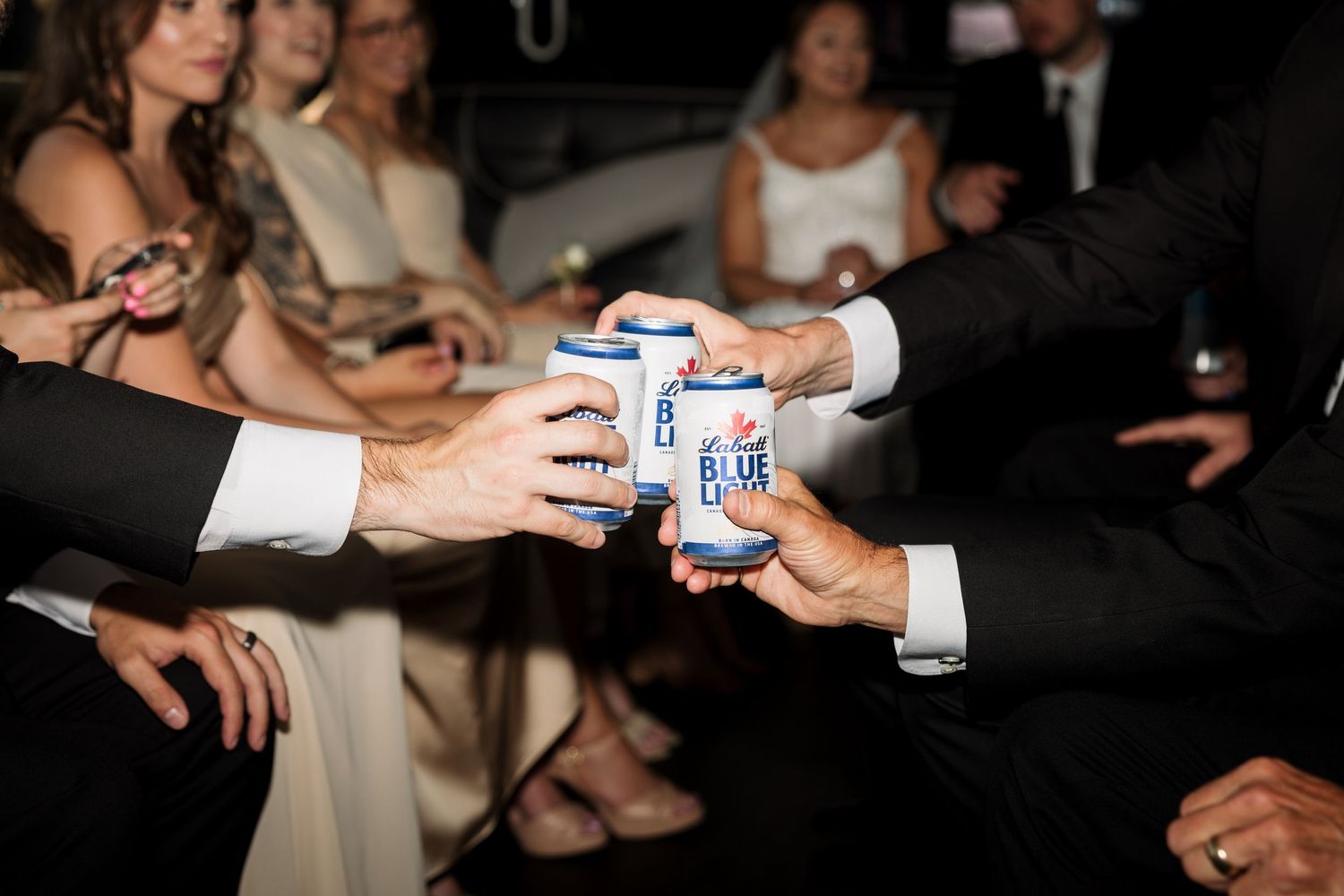 Groomsmen in black tuxedos toasting with Bud Light cans at a wedding reception celebration.