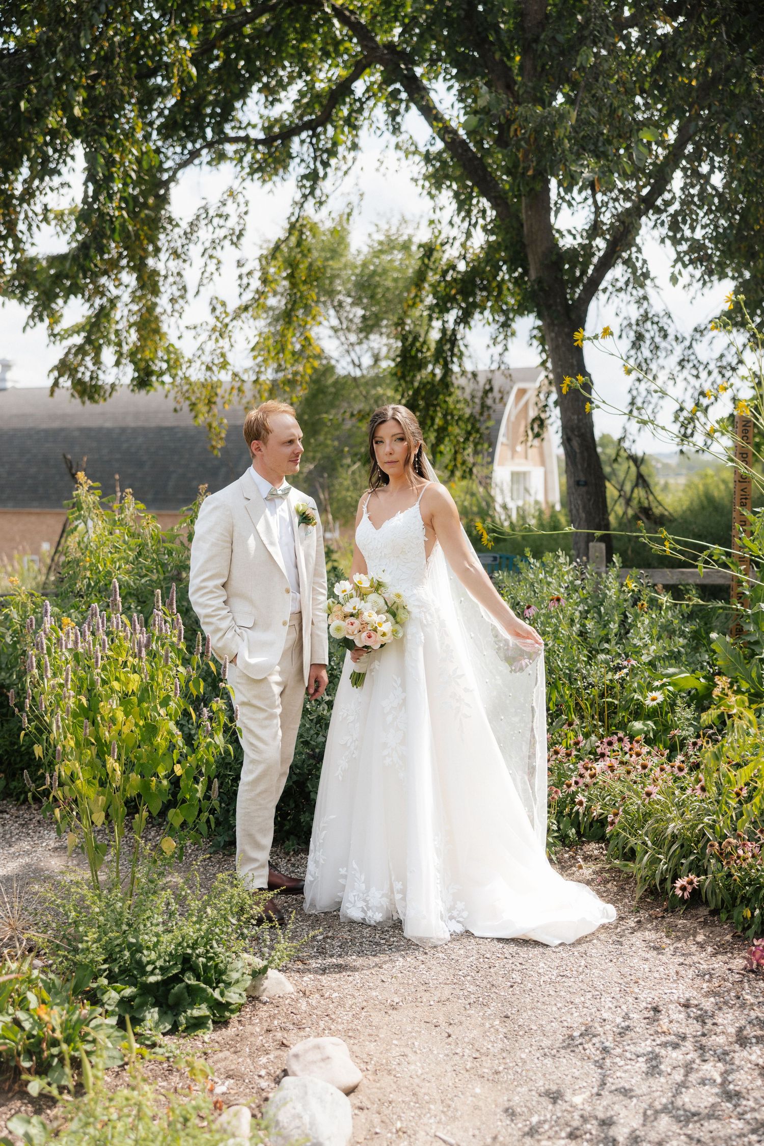 Bride and groom stand together in a lush garden, she holds a bouquet in a white gown, he wears a cream suit.