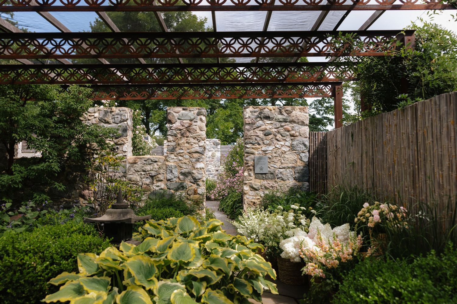 Lush garden with stone wall, wooden pergola, hosta plants, and decorative fountain surrounded by greenery.