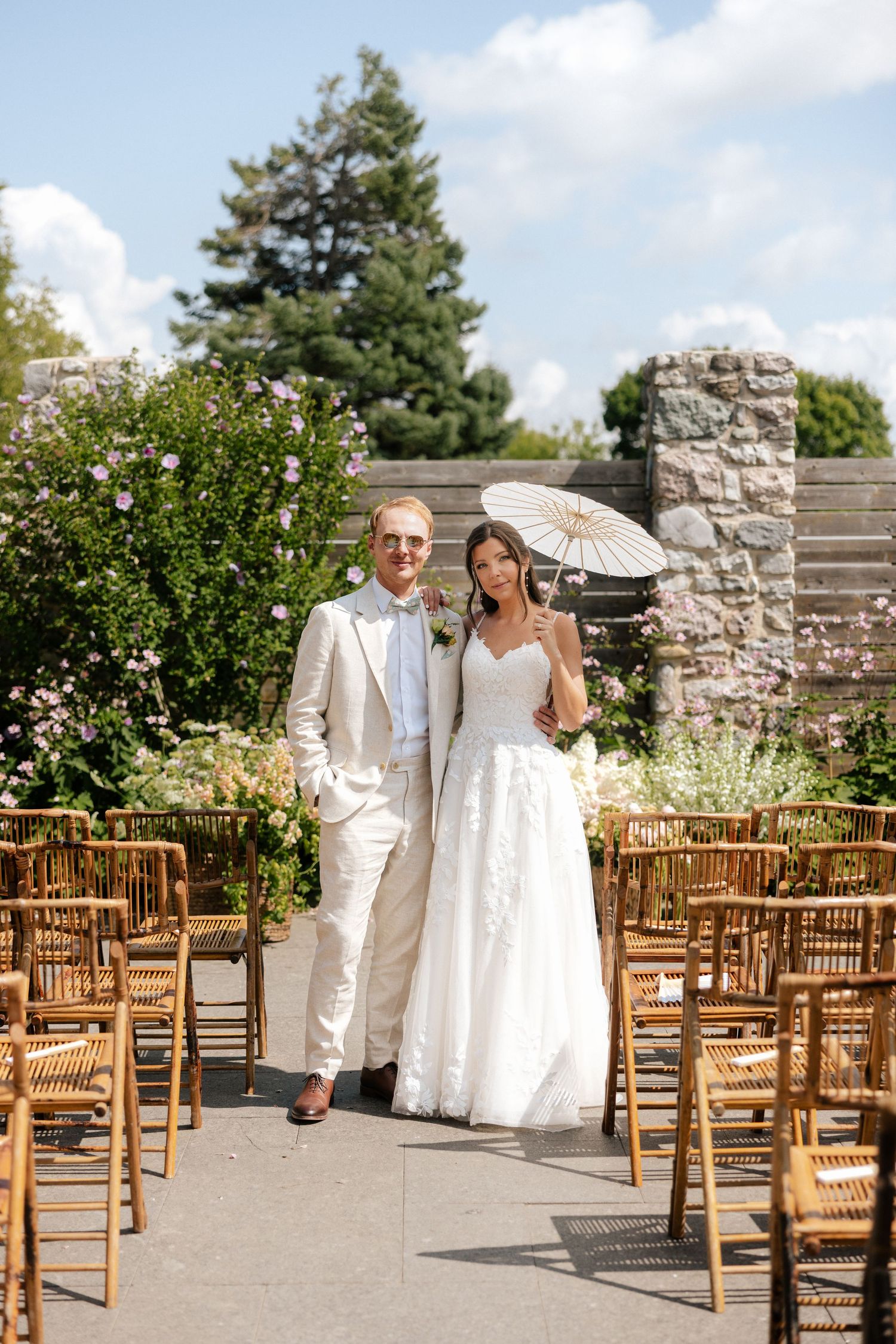 Bride and groom posing in outdoor wedding ceremony aisle with parasol and bamboo chairs.