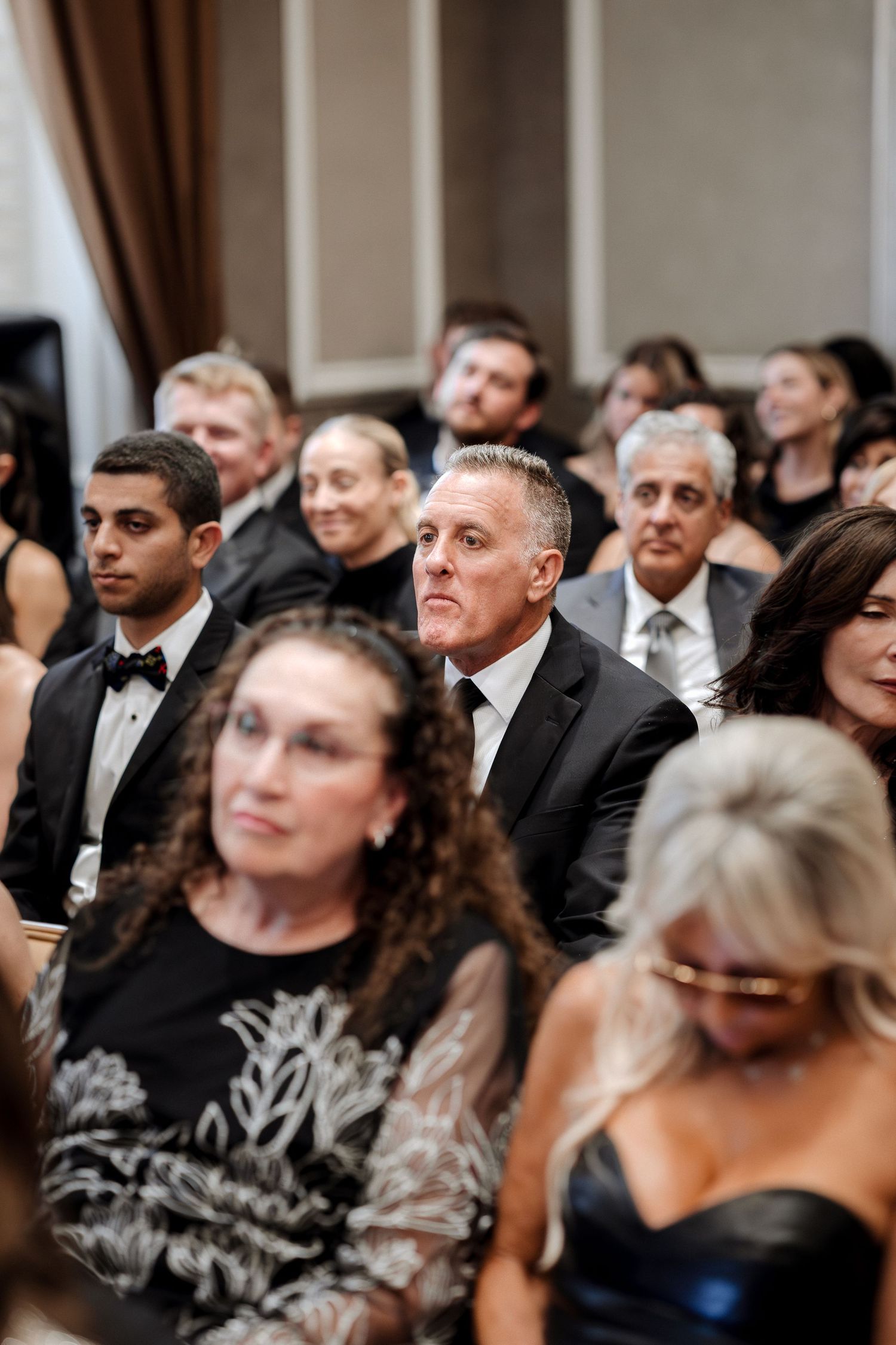 Wedding guests dressed in formal black attire sit attentively during an indoor wedding ceremony.