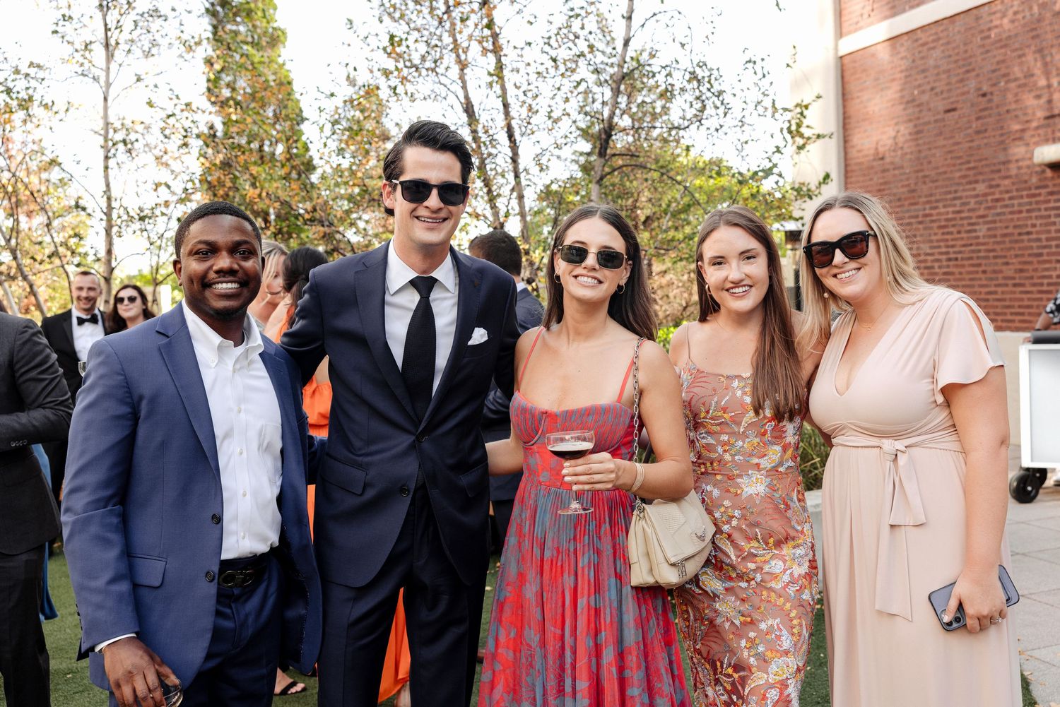 Five young adults smiling together at an outdoor event, dressed in formal attire on a sunny day.