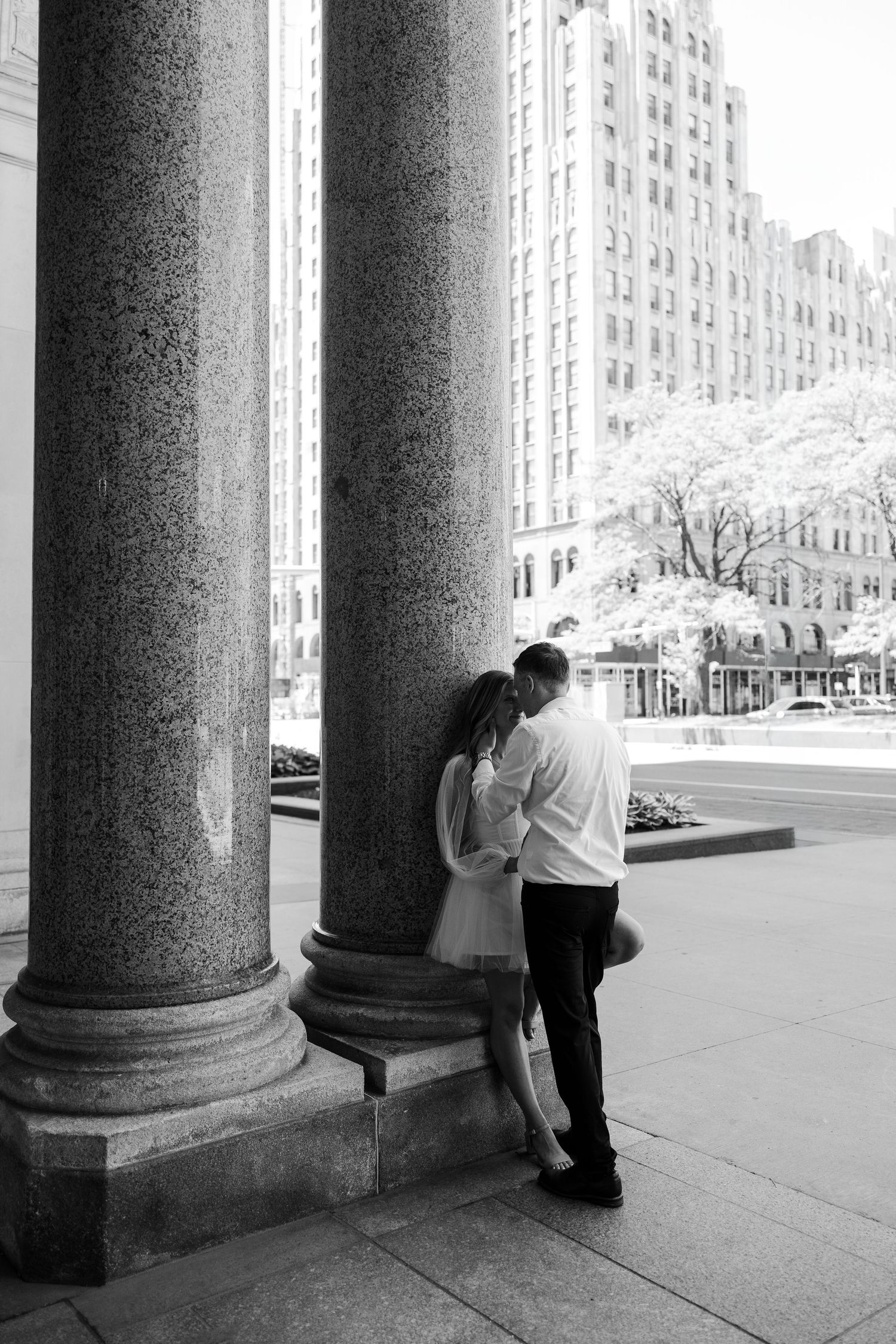Black and white photo of a couple sharing a romantic moment between grand stone columns in an urban cityscape.