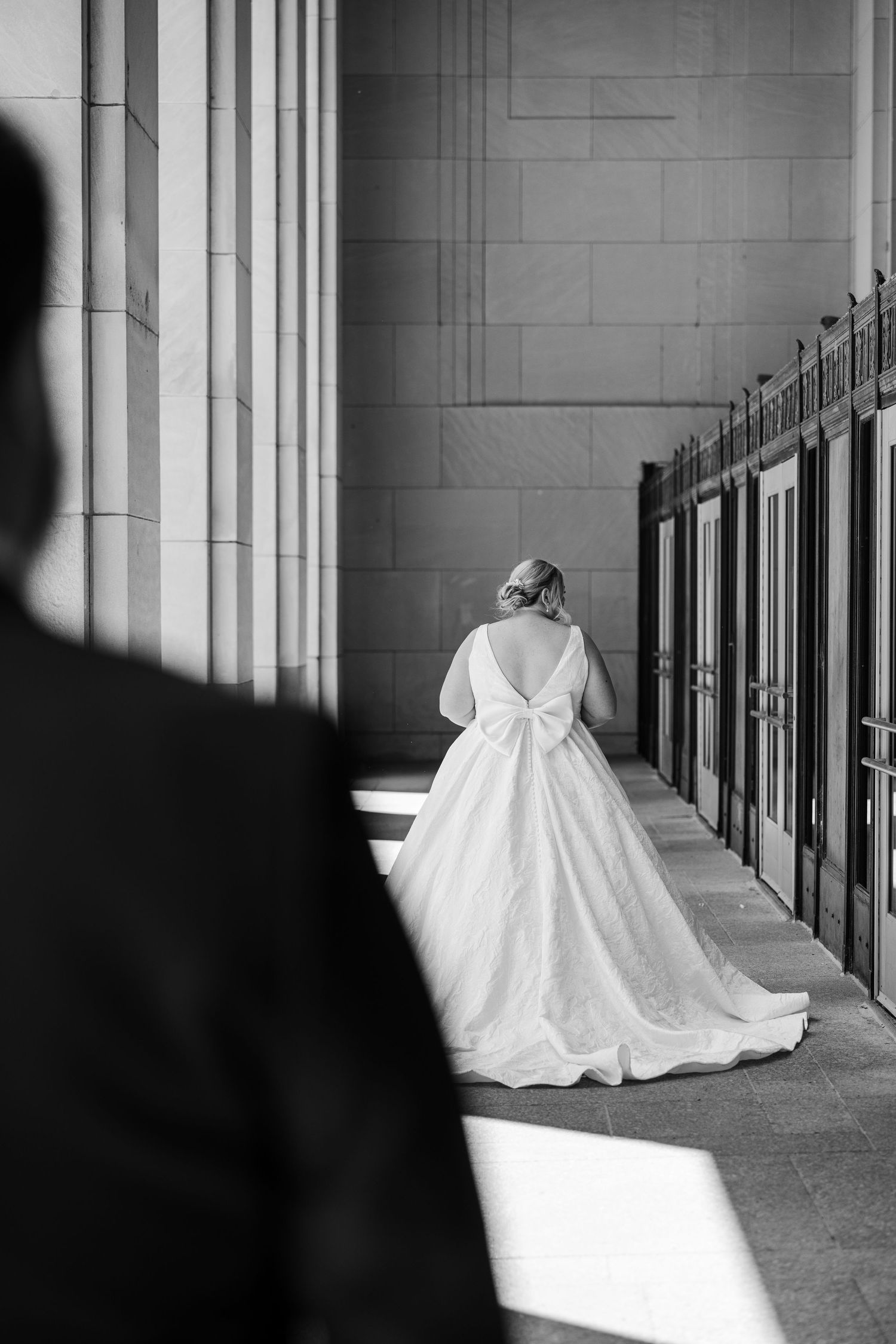 Black and white photo of a bride in a ballgown with bow back detail walking down a grand columned hallway.