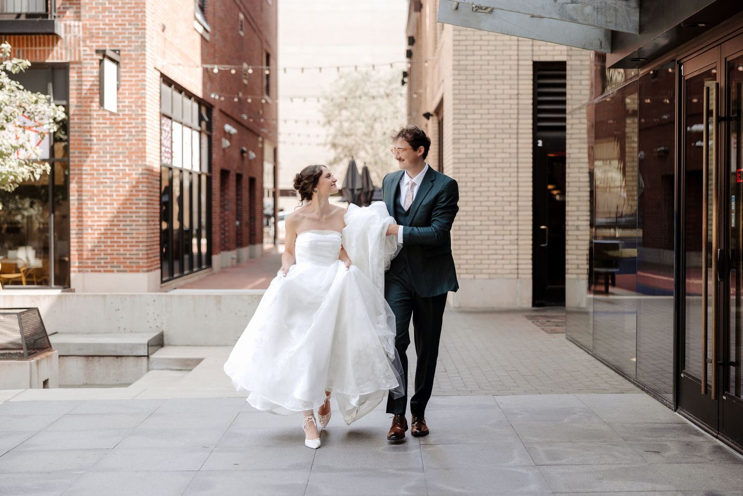 Bride in a white strapless gown and groom in a dark suit walk joyfully together along an urban pathway.
