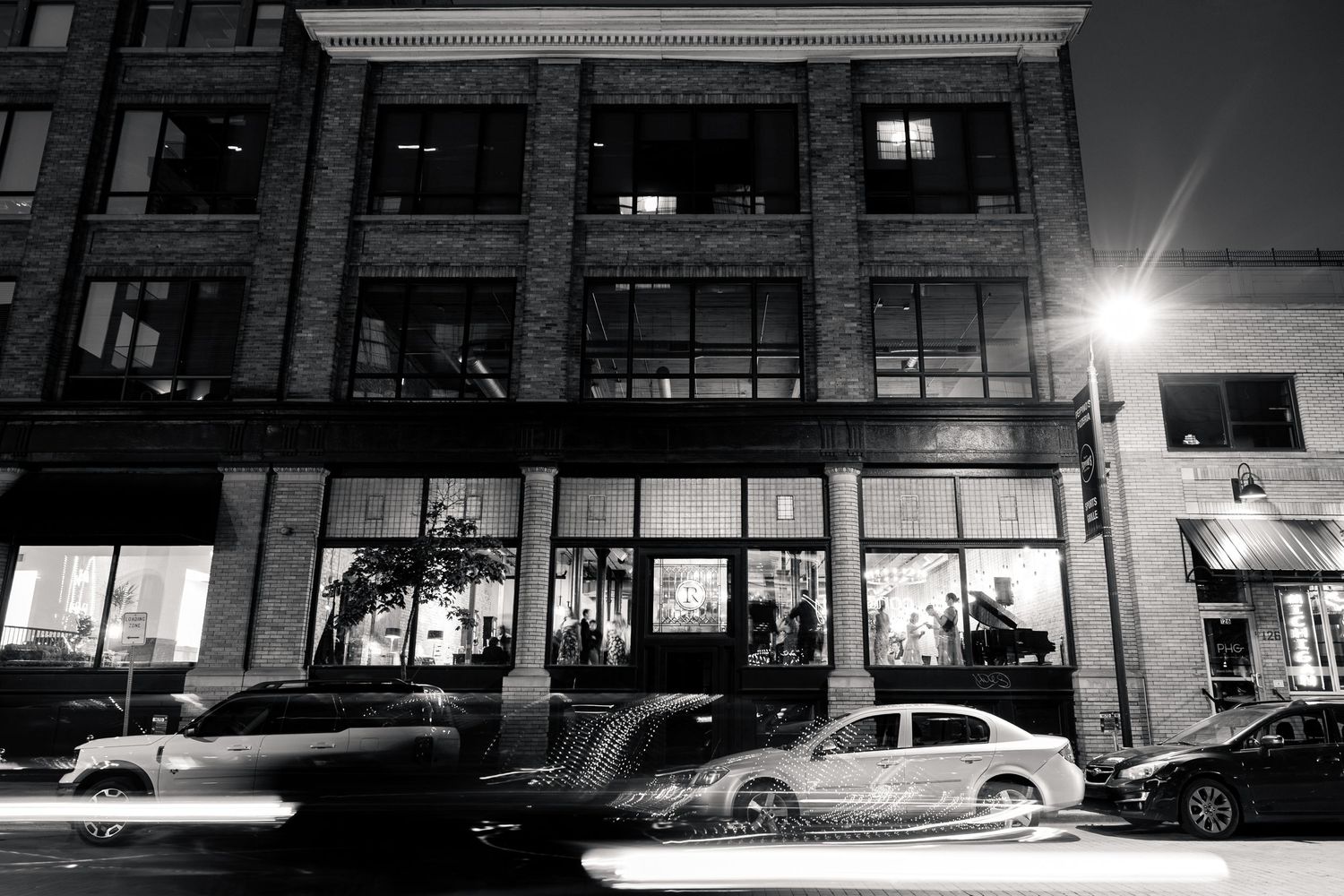 Nighttime exterior of a brick building with illuminated storefronts and passing cars, black and white.
