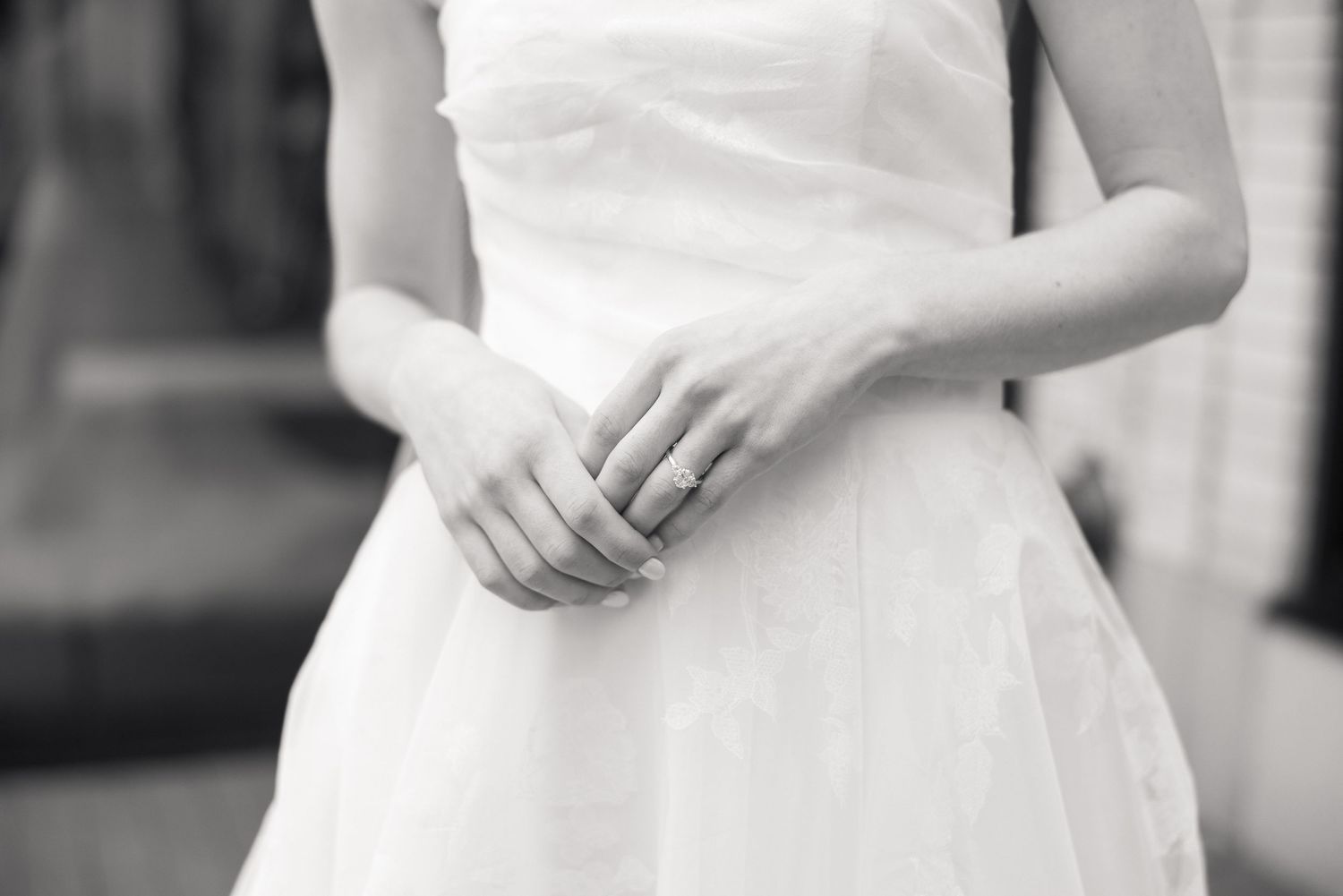 Close-up black and white image of a bride in a white dress with hands folded, showing a delicate ring.