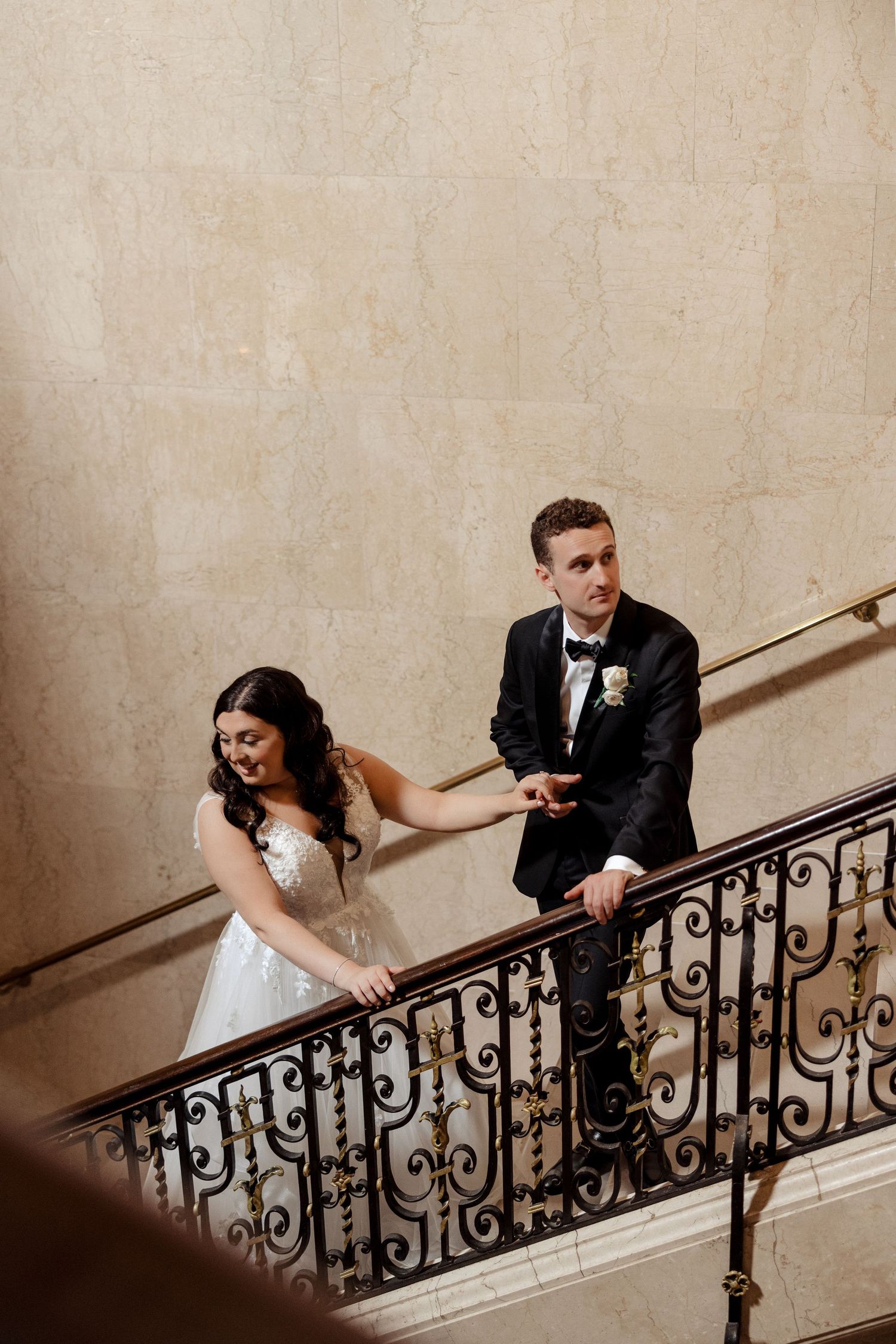 Bride and groom holding hands on an ornate iron staircase in an elegant marble building.