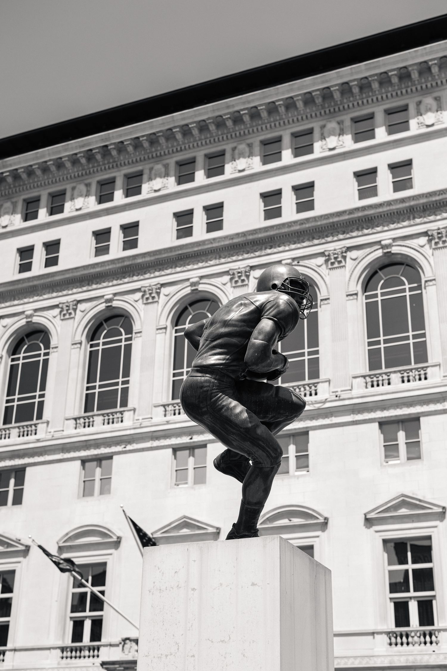 Black and white photo of a bronze football player statue in front of a classical building facade.