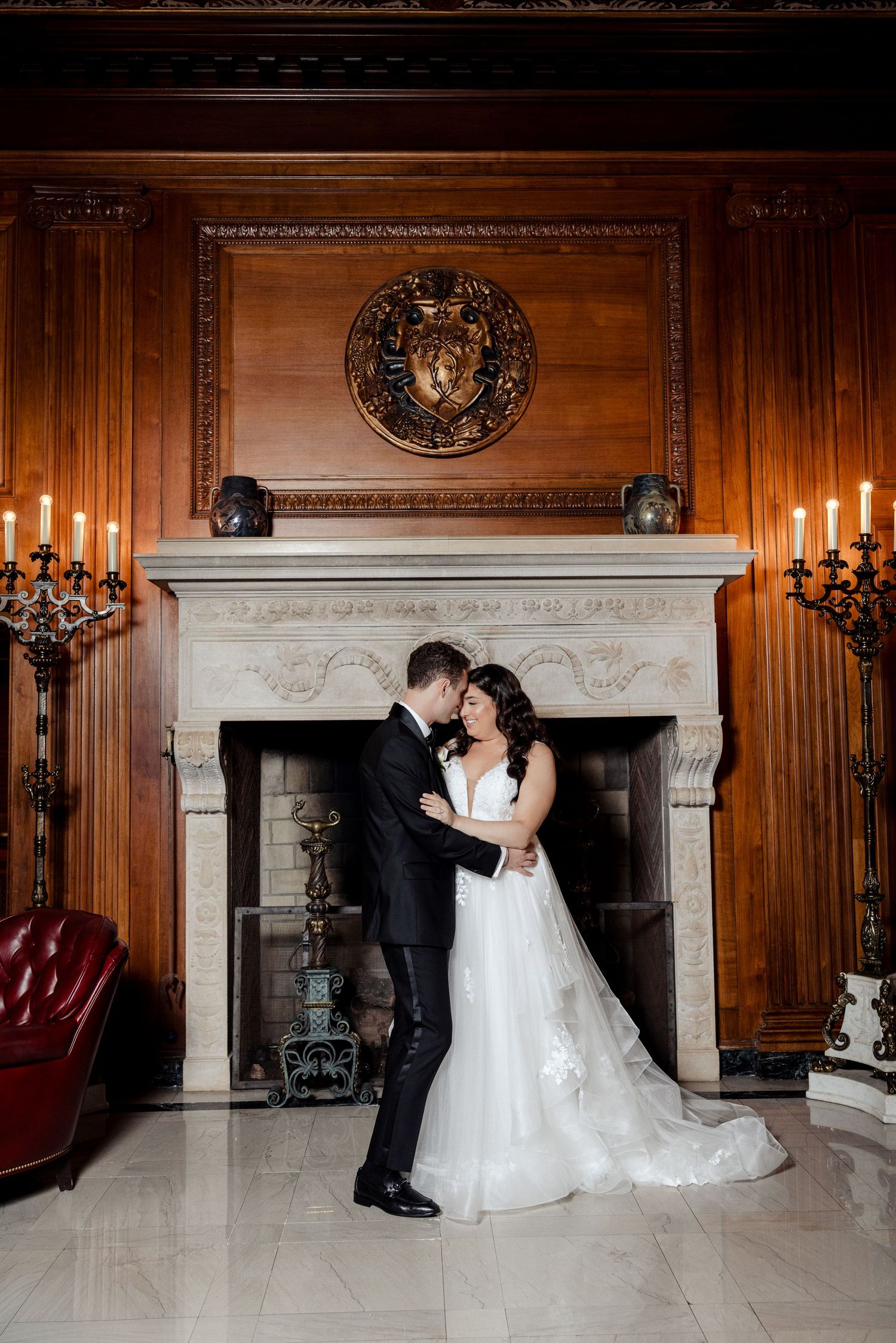 Bride and groom embracing before ornate marble fireplace in candlelit wood-paneled room.