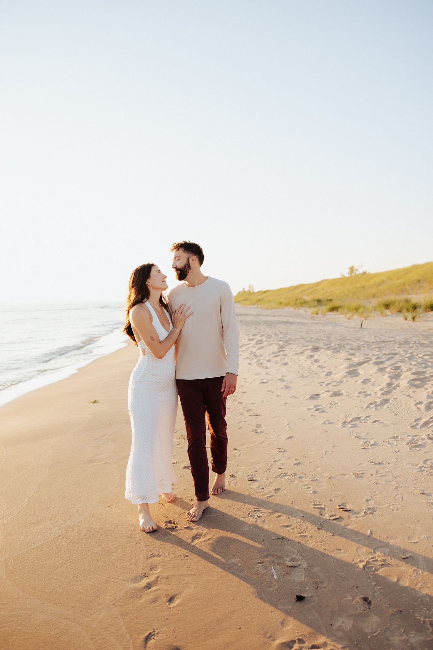 Couple walking hand in hand on a sandy beach at golden hour, woman in white dress, man in cream top and burgundy pants.
