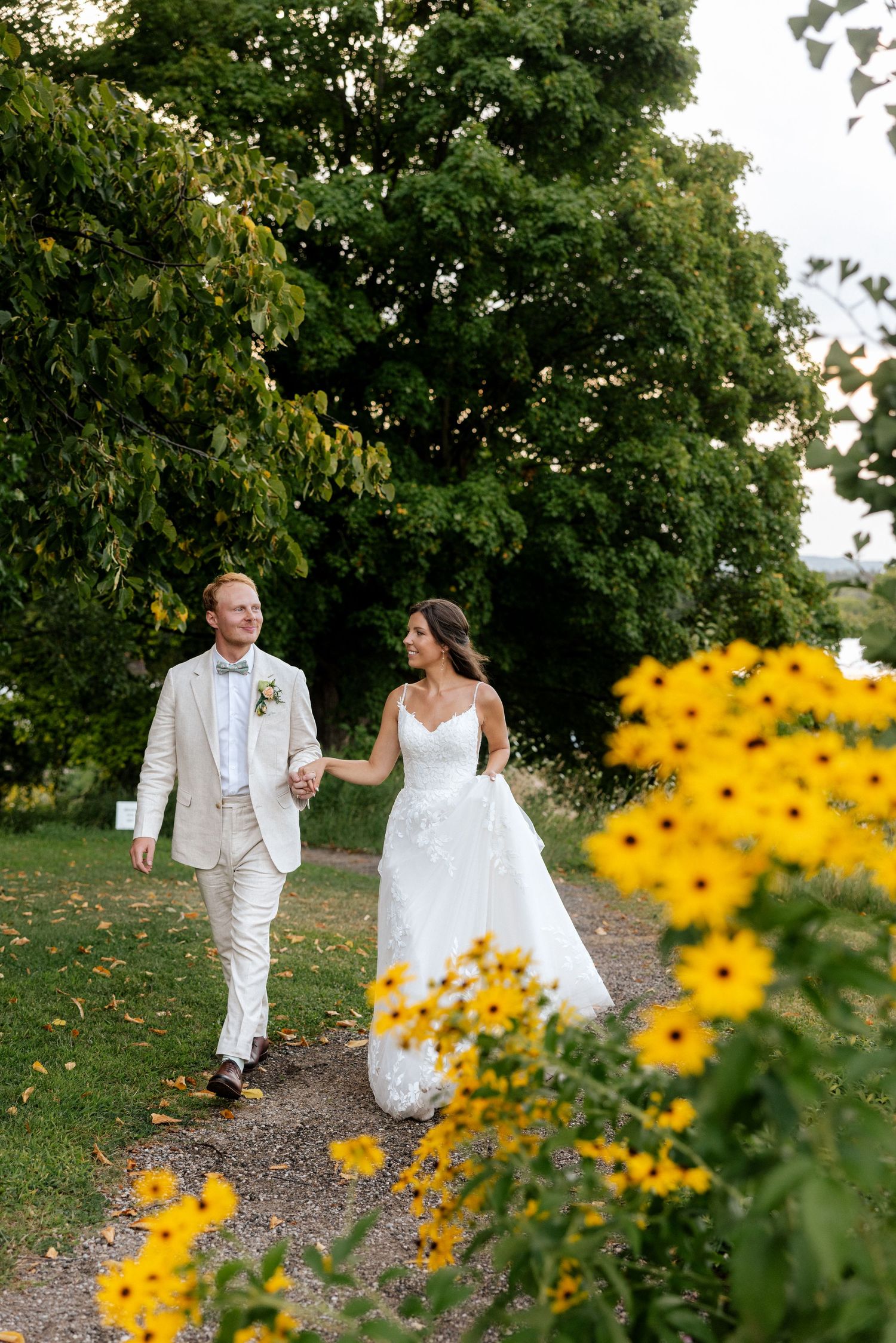 A bride and groom walk hand-in-hand along a garden path surrounded by lush trees and yellow flowers.