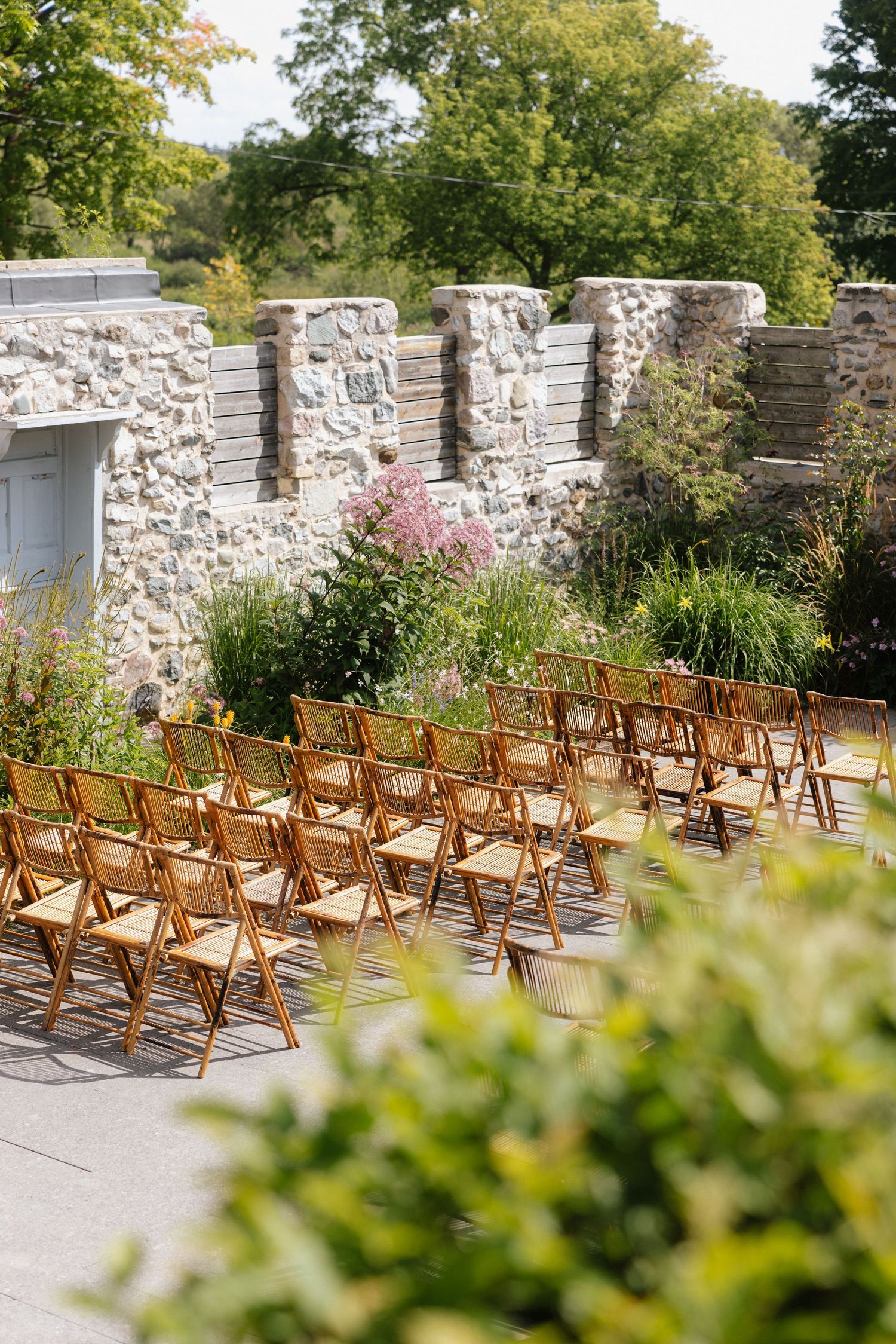 Rows of wooden folding chairs set up for an outdoor wedding ceremony beside a rustic stone wall with lush gardens.