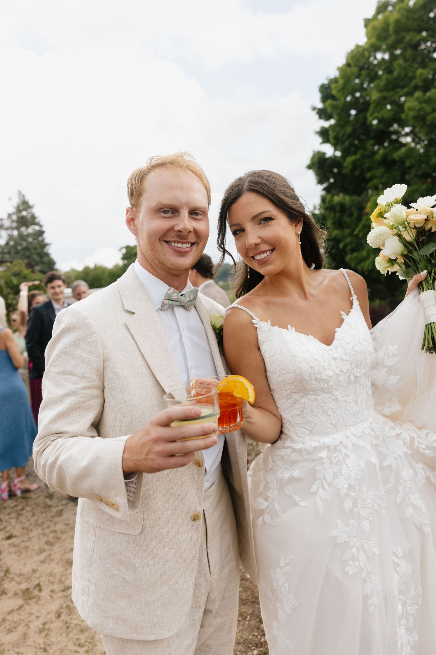 Smiling bride in lace gown and groom in beige suit holding cocktails outdoors at a beach wedding celebration.