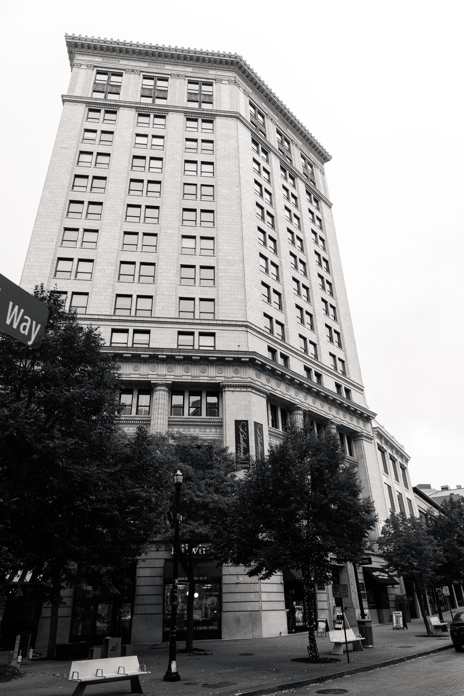 Black and white photo of a tall historic multi-story building on a city street corner with trees and lamp posts.