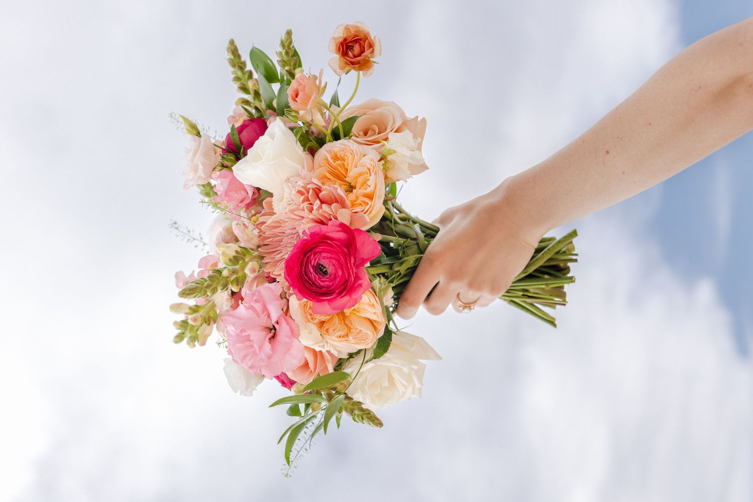 Hand holds a vibrant bridal bouquet of pink, coral, and white flowers against a cloudy sky.