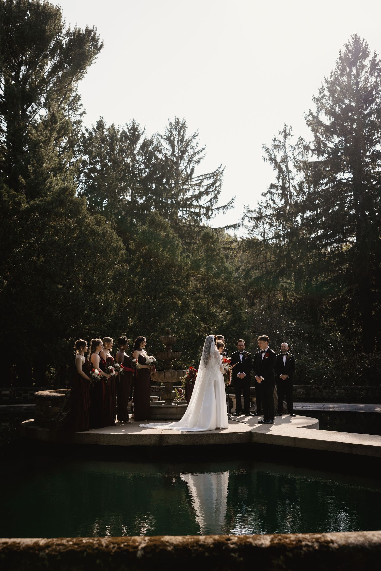 Outdoor wedding ceremony by a reflective pool, bride in white gown with veil, bridesmaids in dark dresses, lush trees backdrop.