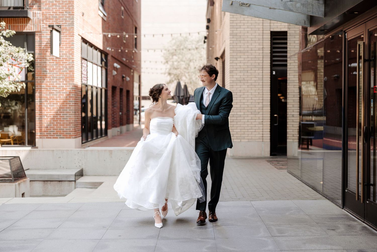 Bride and groom walking hand in hand on a city sidewalk, bride in white gown, groom in dark suit.