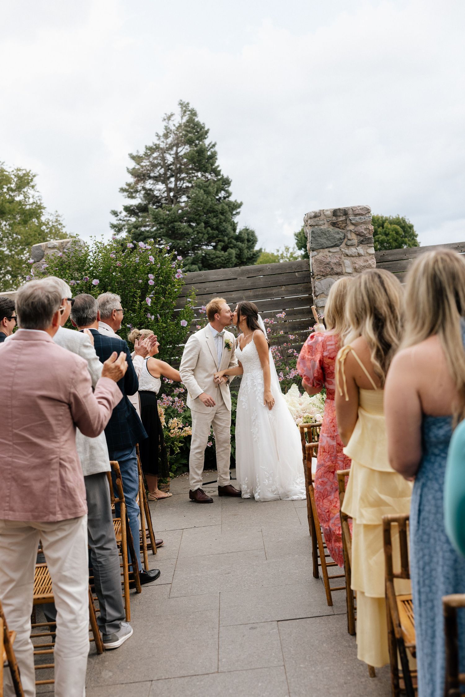 Bride and groom sharing first kiss at outdoor wedding ceremony while guests applaud.