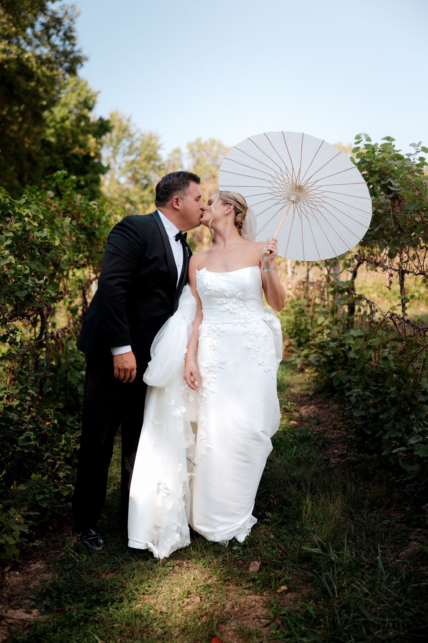 A bride and groom share a kiss outdoors, she holds a white parasol, wearing a strapless gown, he in a black tuxedo.