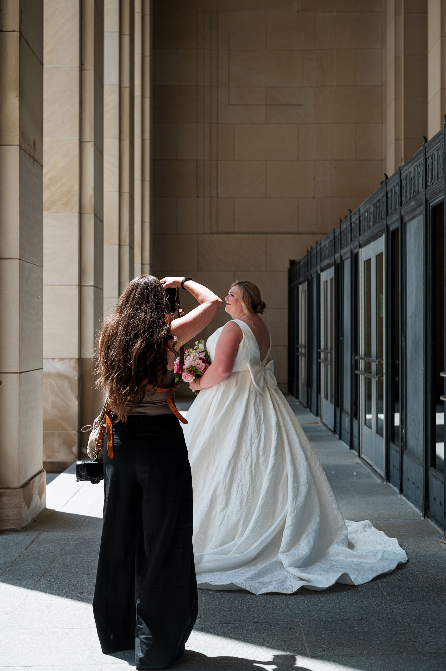 A bridesmaid adjusts a bride's hair outside a grand stone building, as the bride holds a pink floral bouquet.