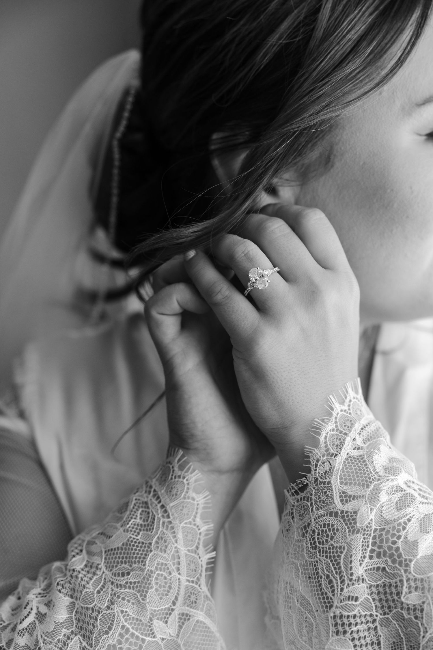 Black and white photo of a bride in lace robe putting on earrings, showcasing her diamond engagement ring.