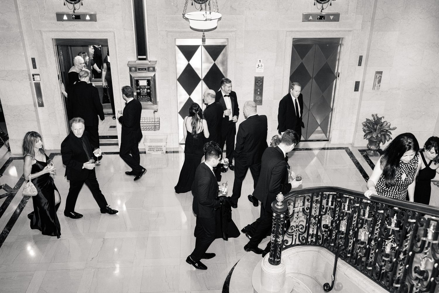 Black and white photo of wedding guests descending grand staircase in formal attire at elegant venue.