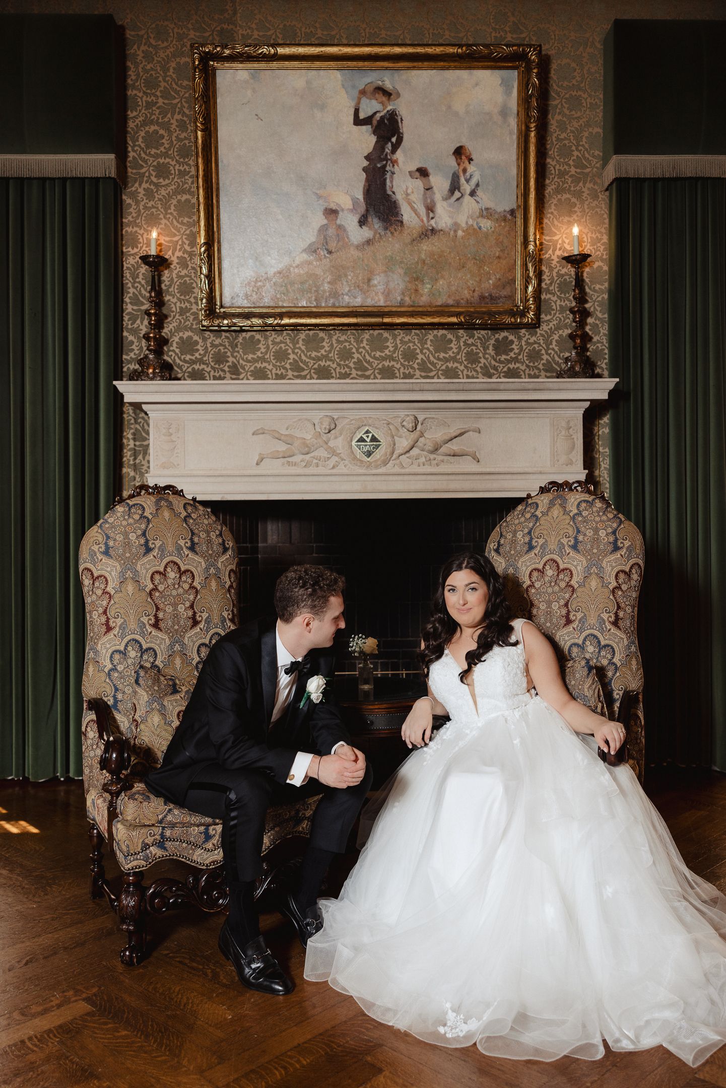Bride and groom sit in ornate chairs by a fireplace, with a classical painting above, in an elegant wedding portrait.
