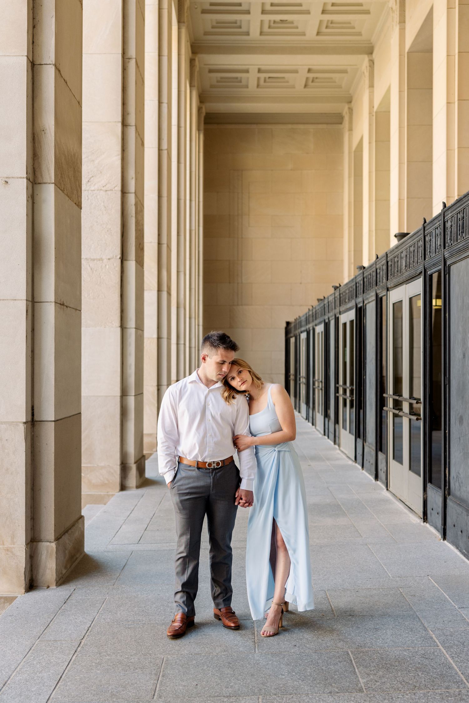 Romantic couple embracing in elegant columned hallway, woman in light blue dress leans on man.