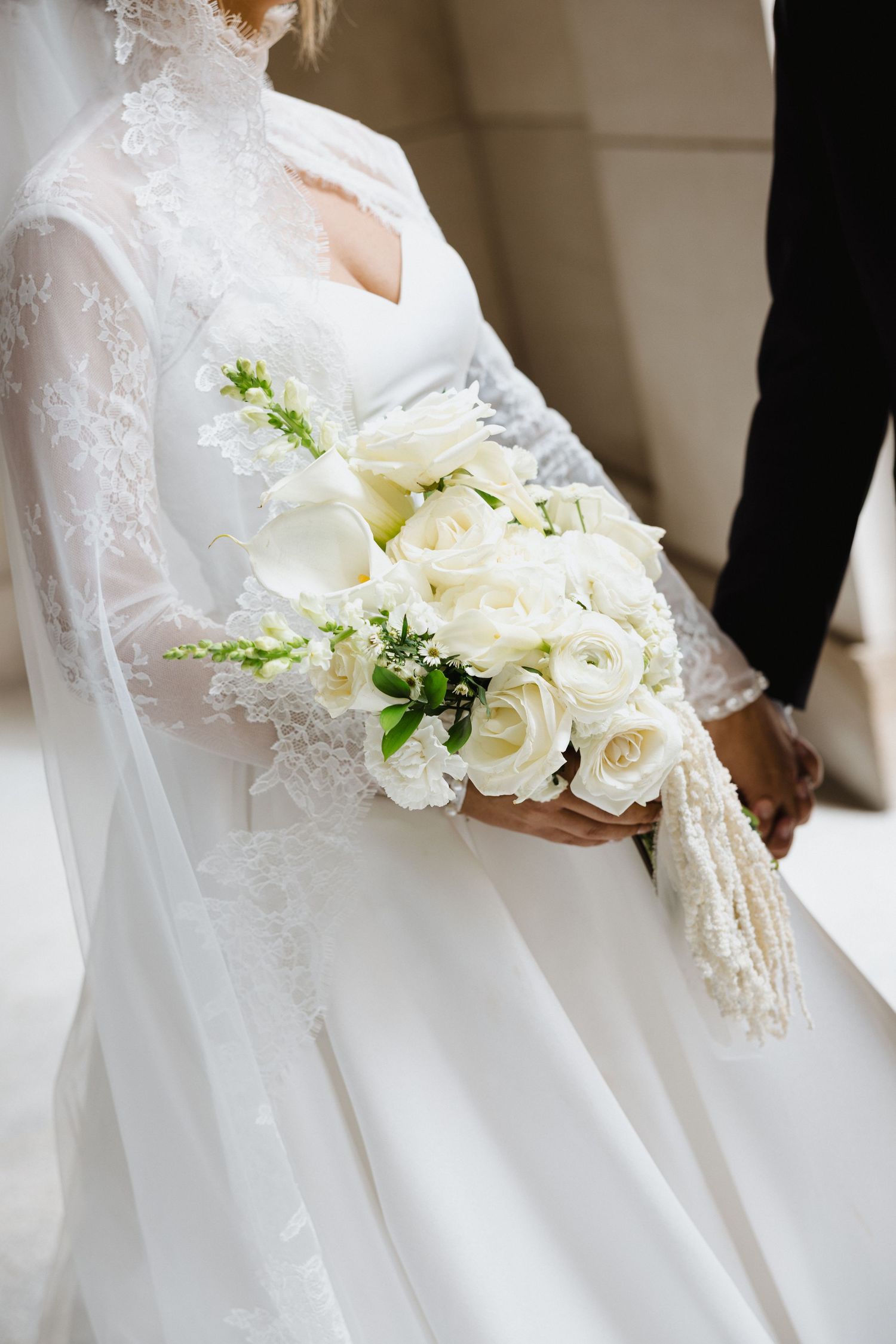 Bride in lace wedding dress holding elegant white rose and calla lily bouquet with greenery accents.