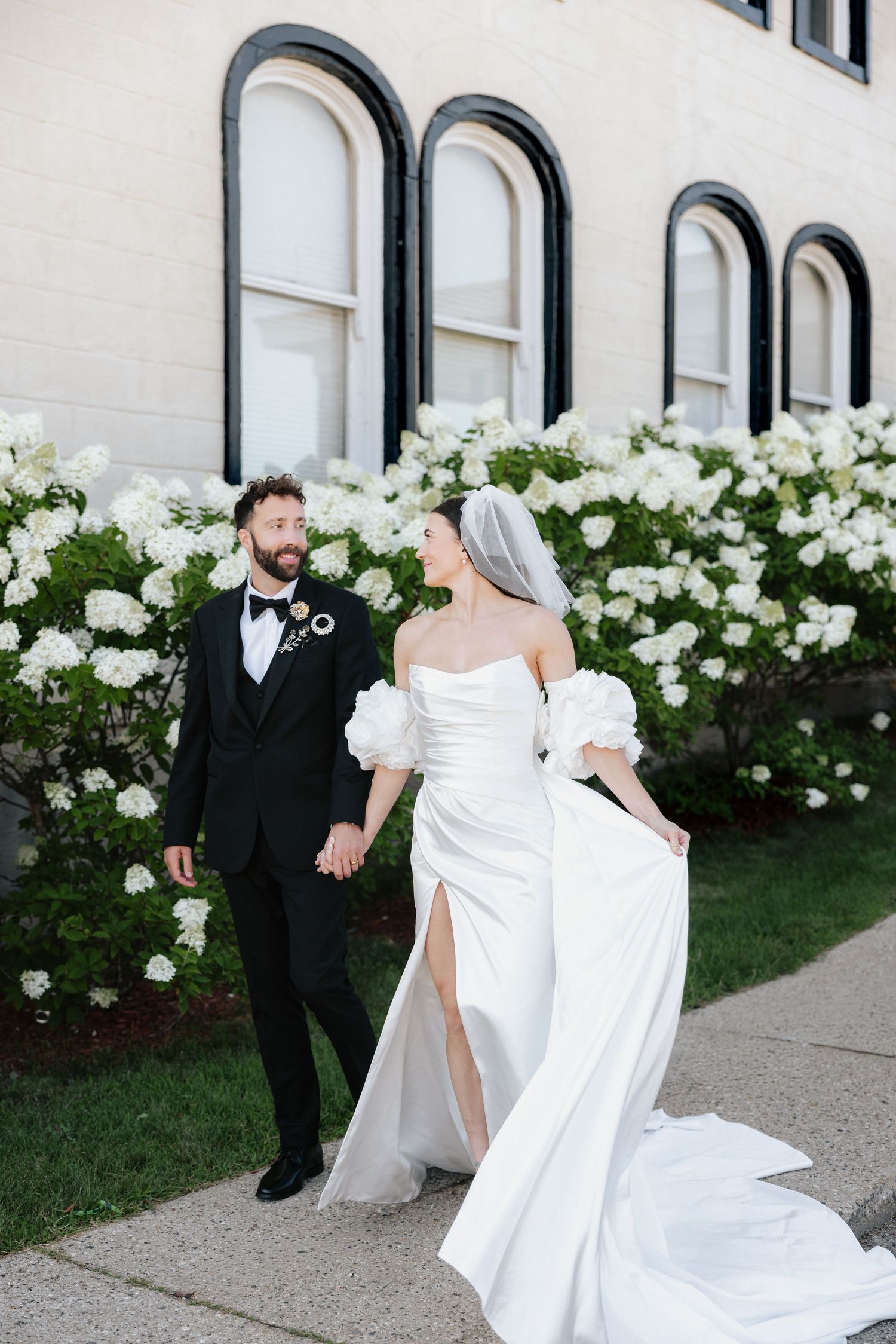 A bride and groom walk hand-in-hand in front of white hydrangea bushes, she in a strapless gown, he in a black tuxedo.